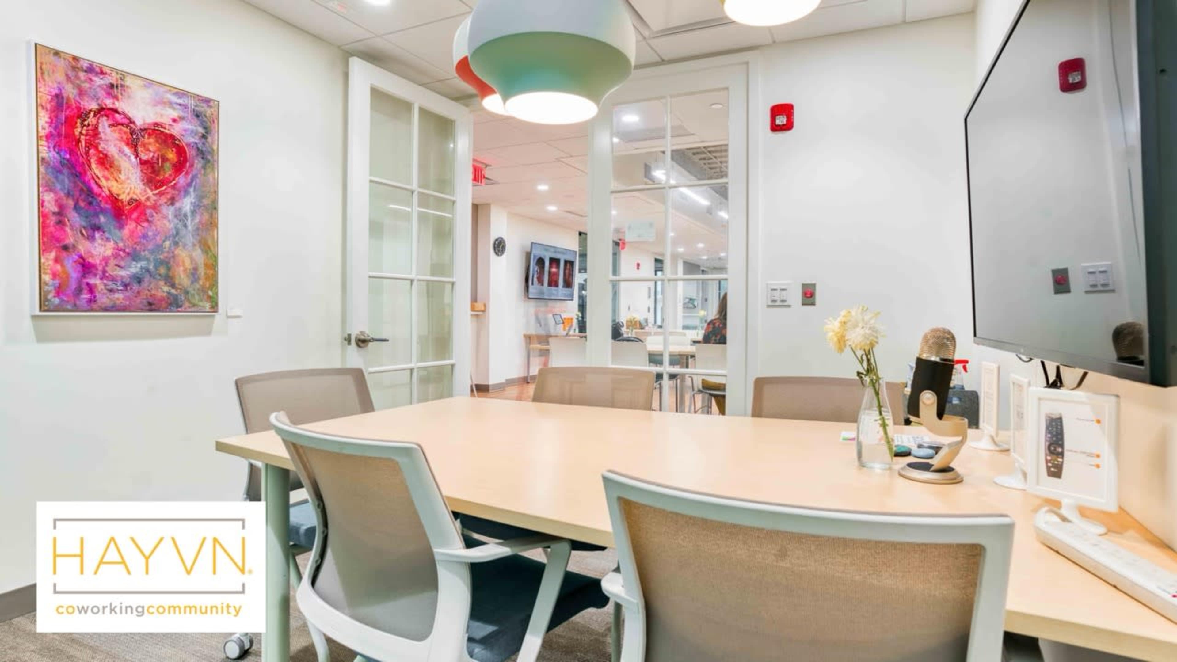 The image shows a bright, modern conference room with a large table, chairs, and a wall-mounted monitor, featuring glass doors and a colorful artwork.