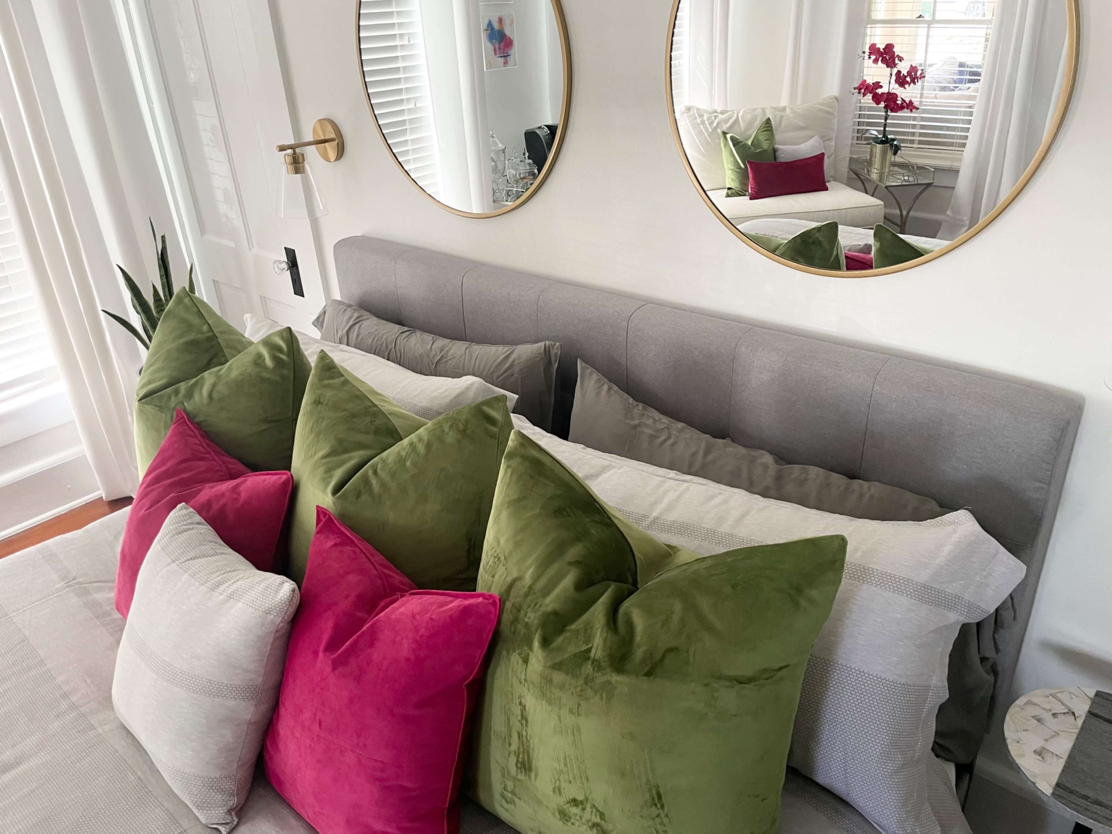 The image shows a bed with a collection of colorful pillows arranged on a gray headboard, along with two round mirrors reflecting the room's decor.