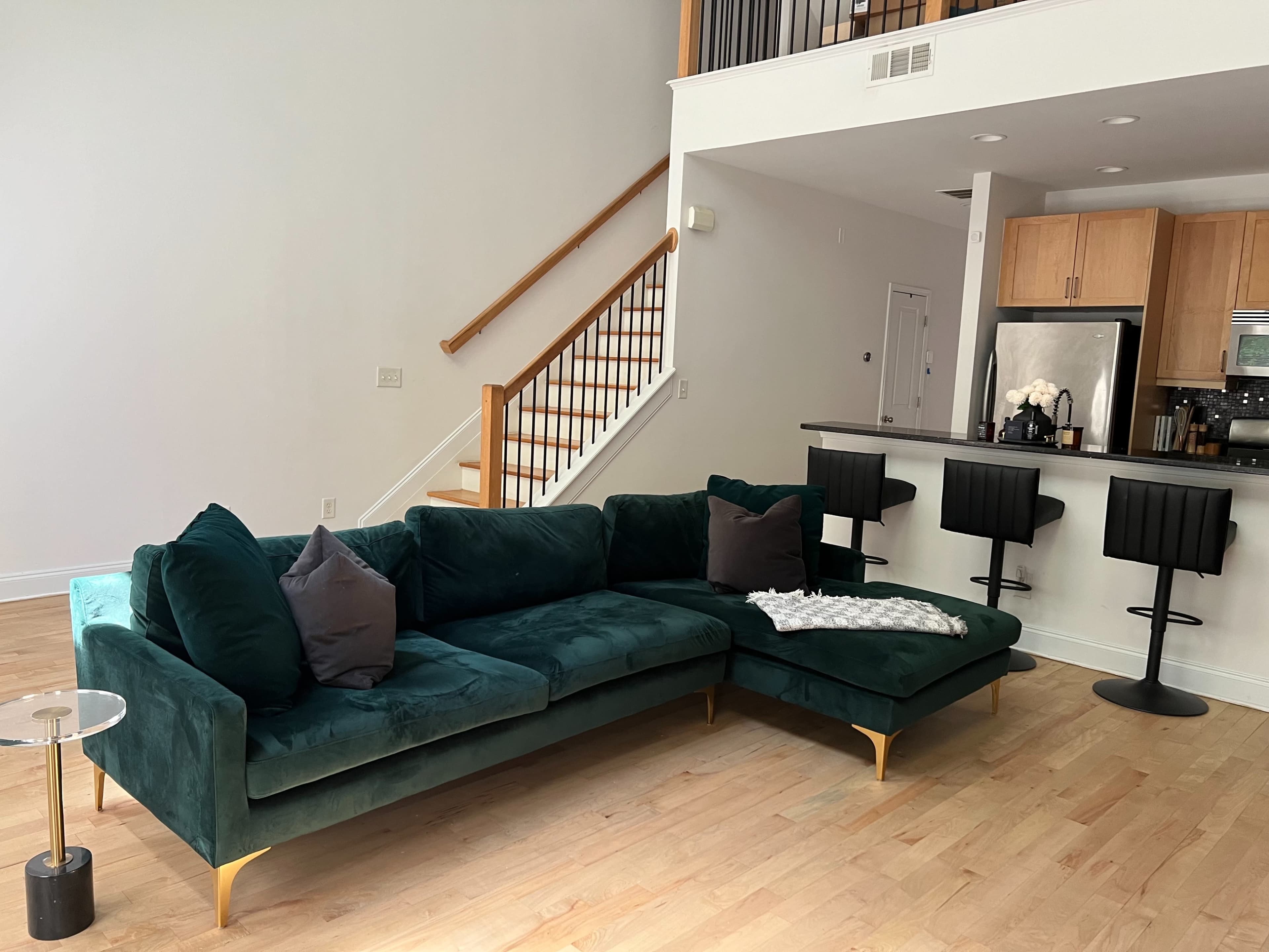 A modern living room features a large green sectional sofa positioned in front of a kitchen with wooden cabinets and black bar stools.
