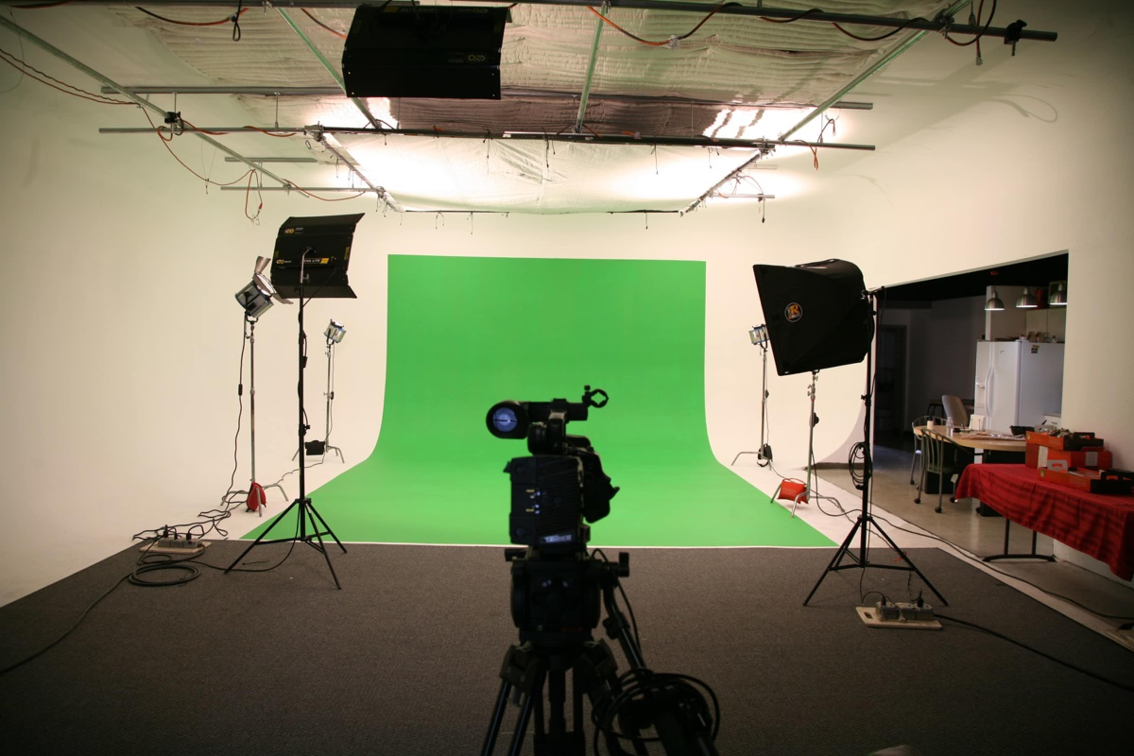 The image shows a photography studio with a green backdrop, a camera on a tripod, and studio lights positioned around the setup.