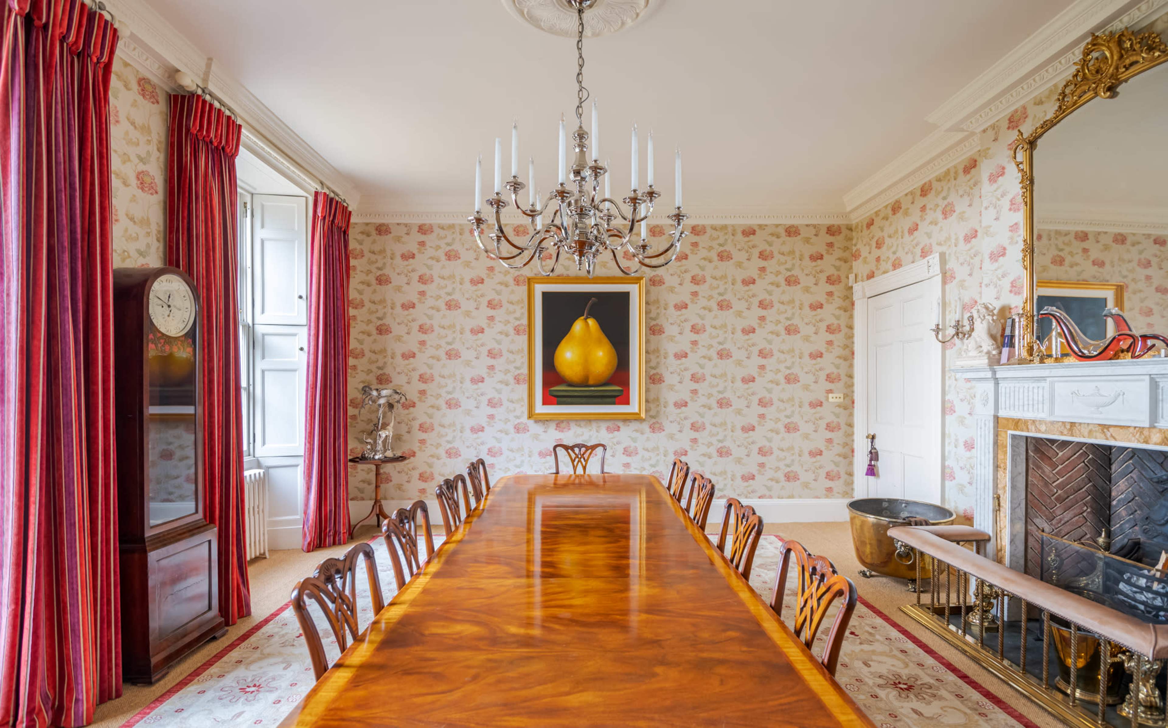 A long wooden dining table is set in an elegantly decorated room with floral wallpaper, framed artwork, and a chandelier hanging above.