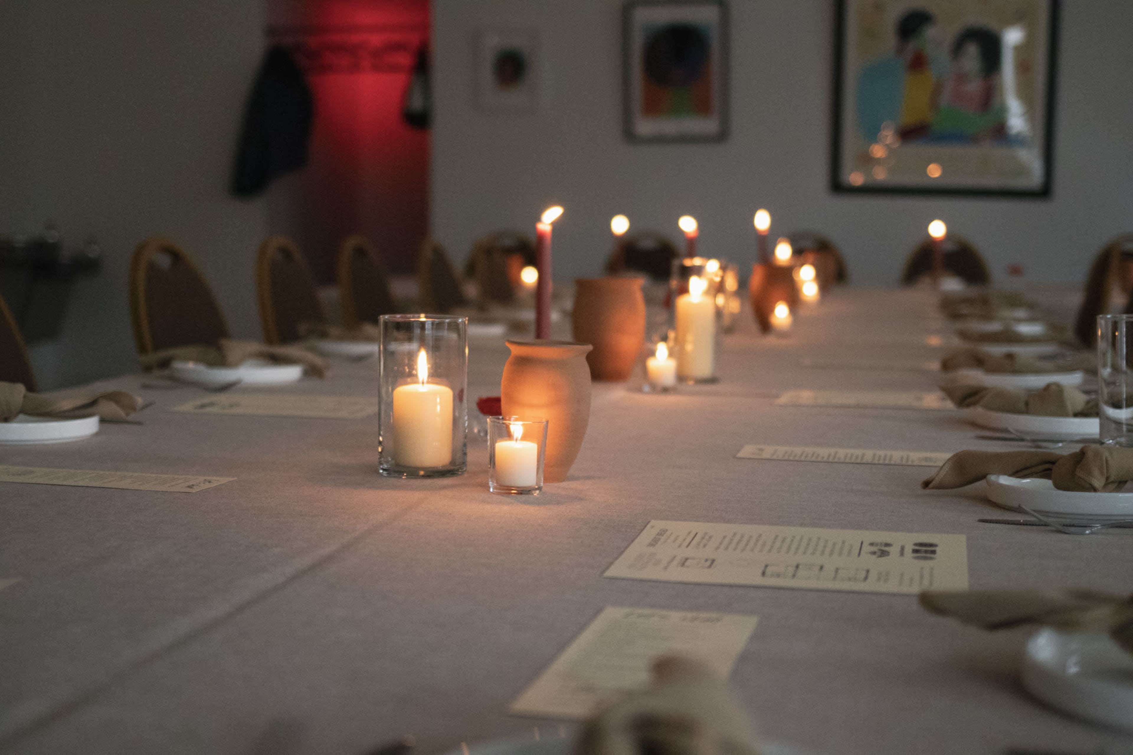 A long dining table is set with candles, glass holders, and a clay pot, creating a warm and inviting atmosphere for a meal.