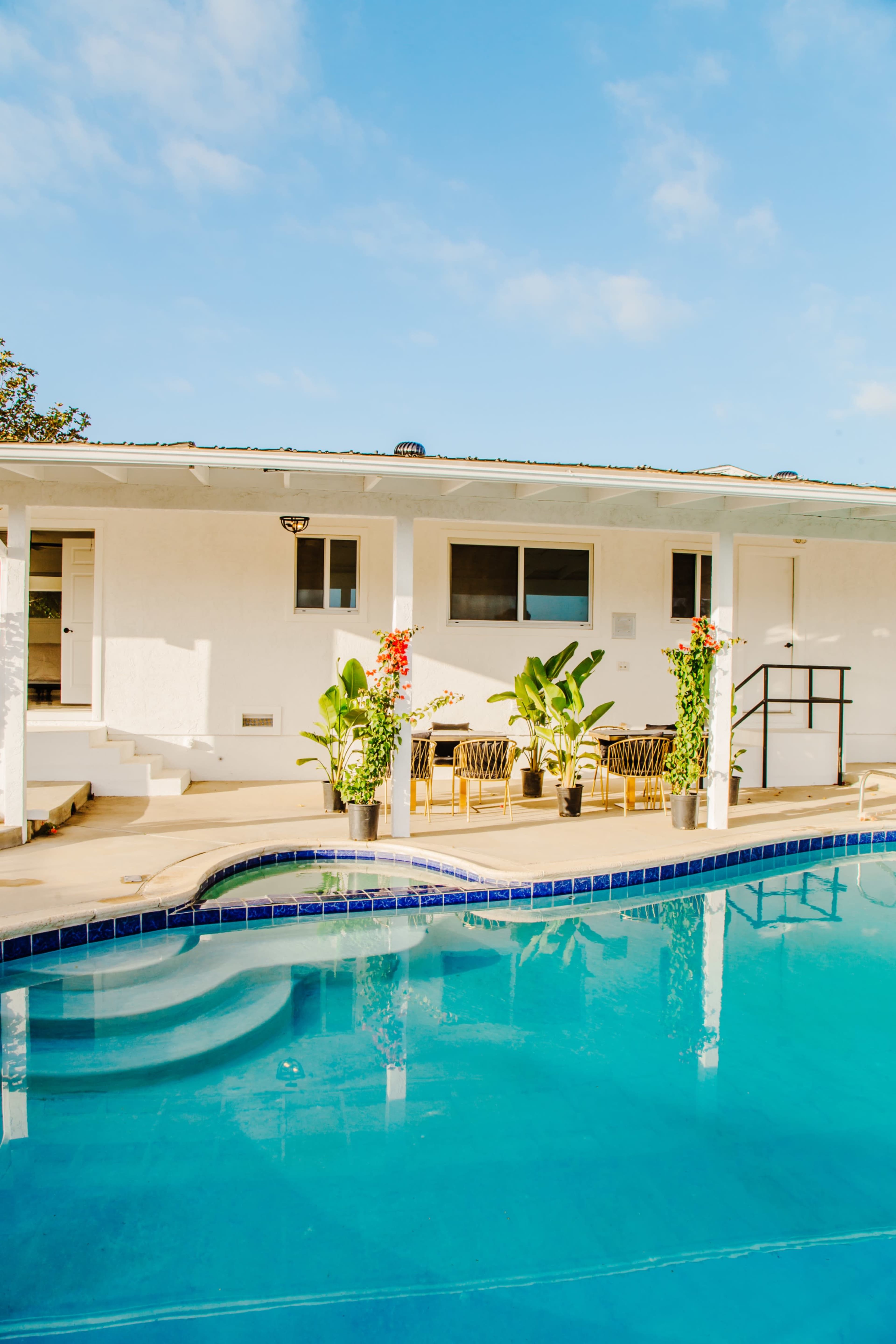A clear blue swimming pool reflects a white house with outdoor seating and potted plants nearby.
