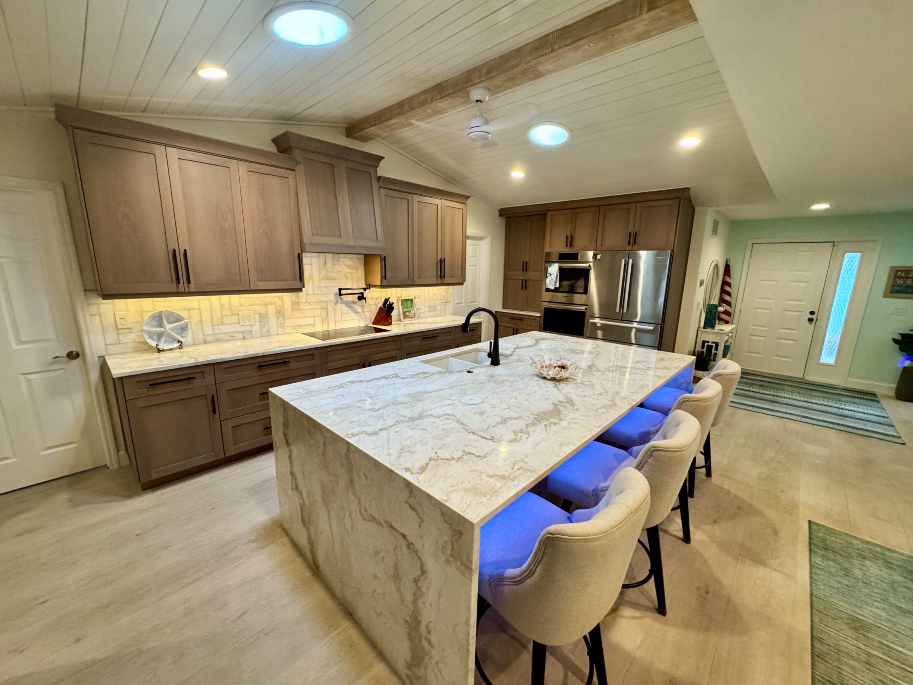 The image shows a modern kitchen featuring wooden cabinetry, a large stone island with bar seating, and recessed lighting.