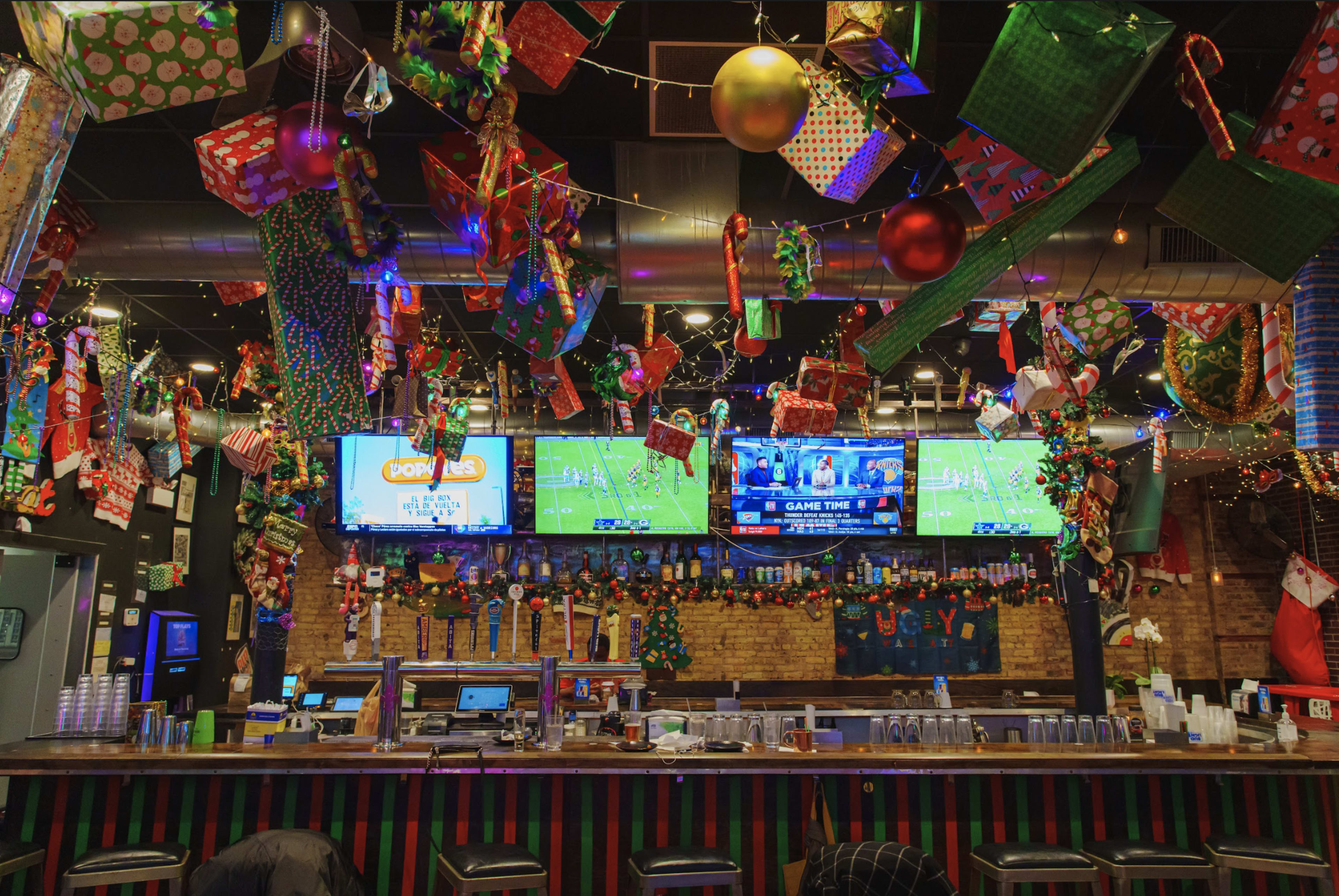 A festive bar is decorated with holiday ornaments, lights, and multiple screens displaying a sports game.