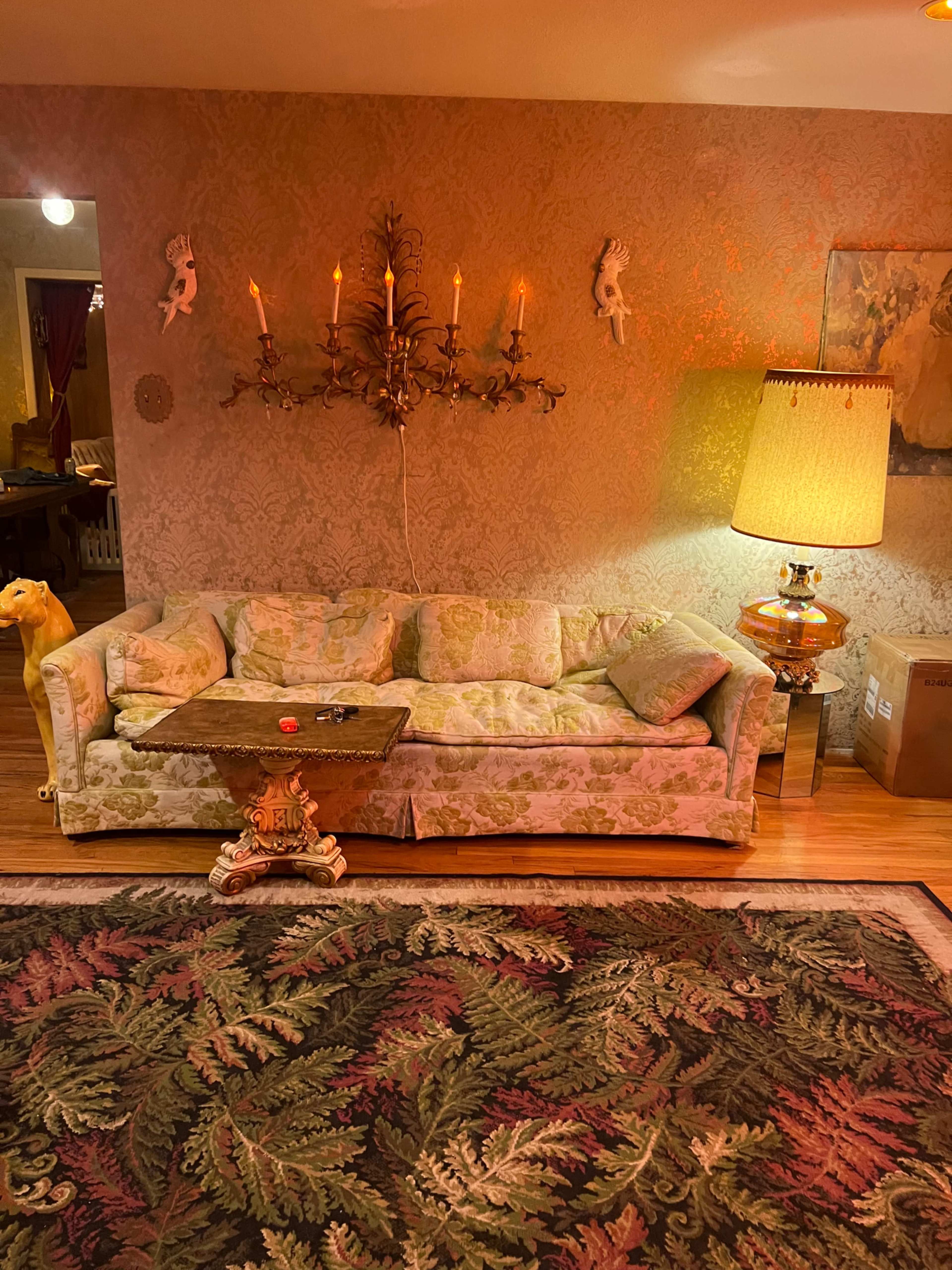 The image shows a vintage floral-patterned couch with a small ornate table and a lamp beside it, situated on a decorative rug in a warmly lit room.
