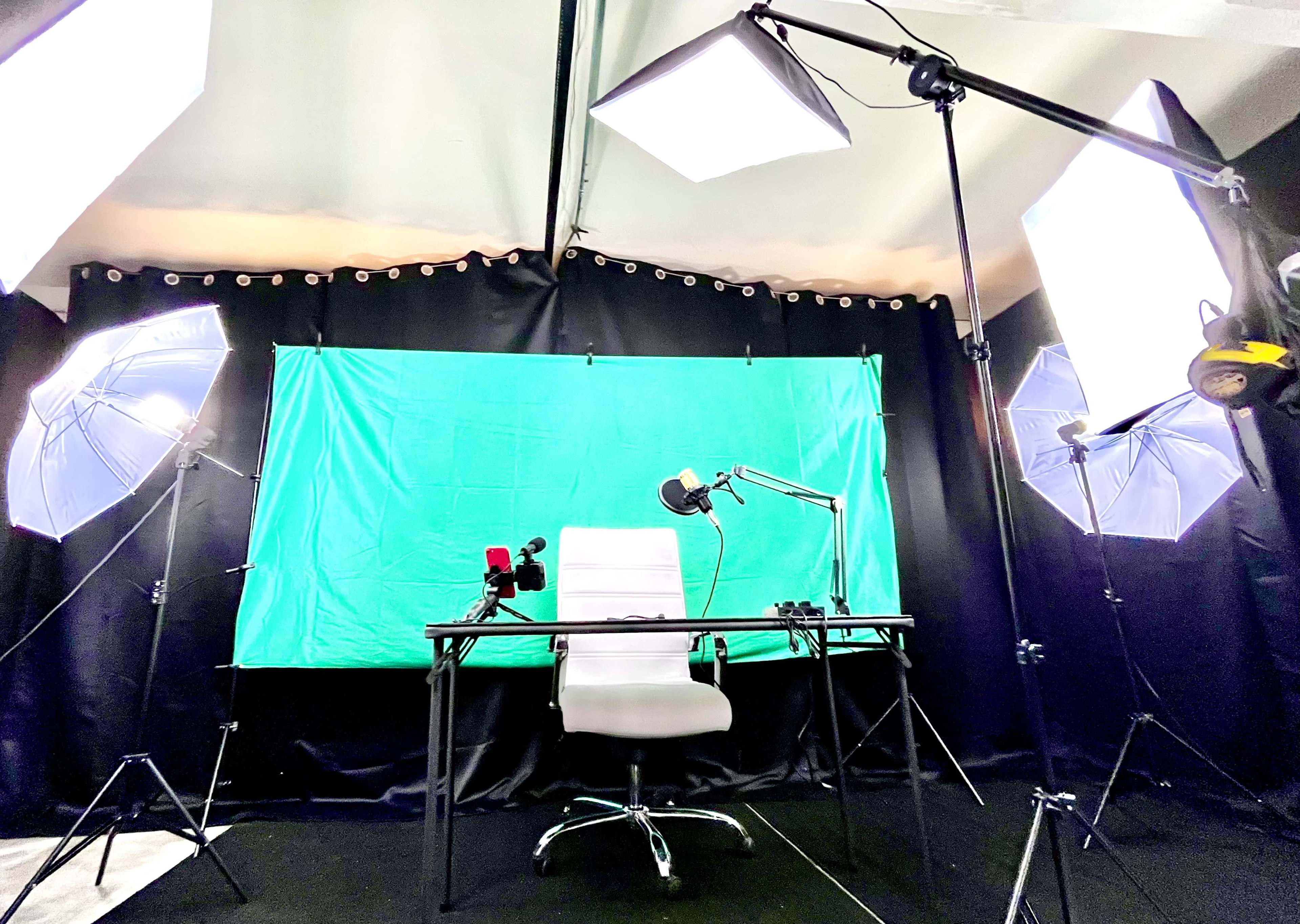 A recording studio setup with a green backdrop, a white chair, a desk, and several lighting devices positioned around the space.