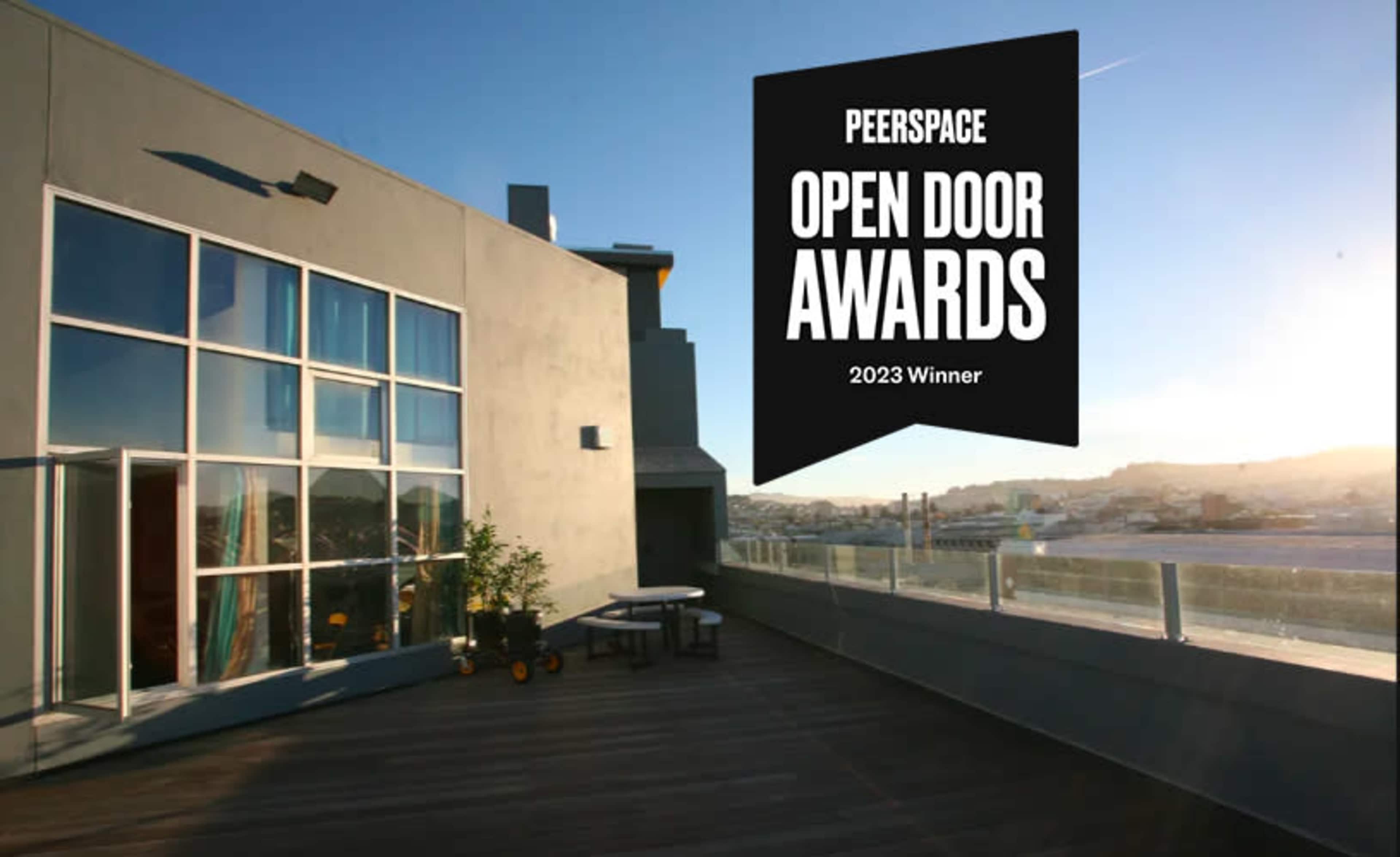 A modern rooftop space with large windows, a wooden deck, and a table set for outdoor seating, highlighted by a banner announcing the Peerspace Open Door Awards 2023 Winner.