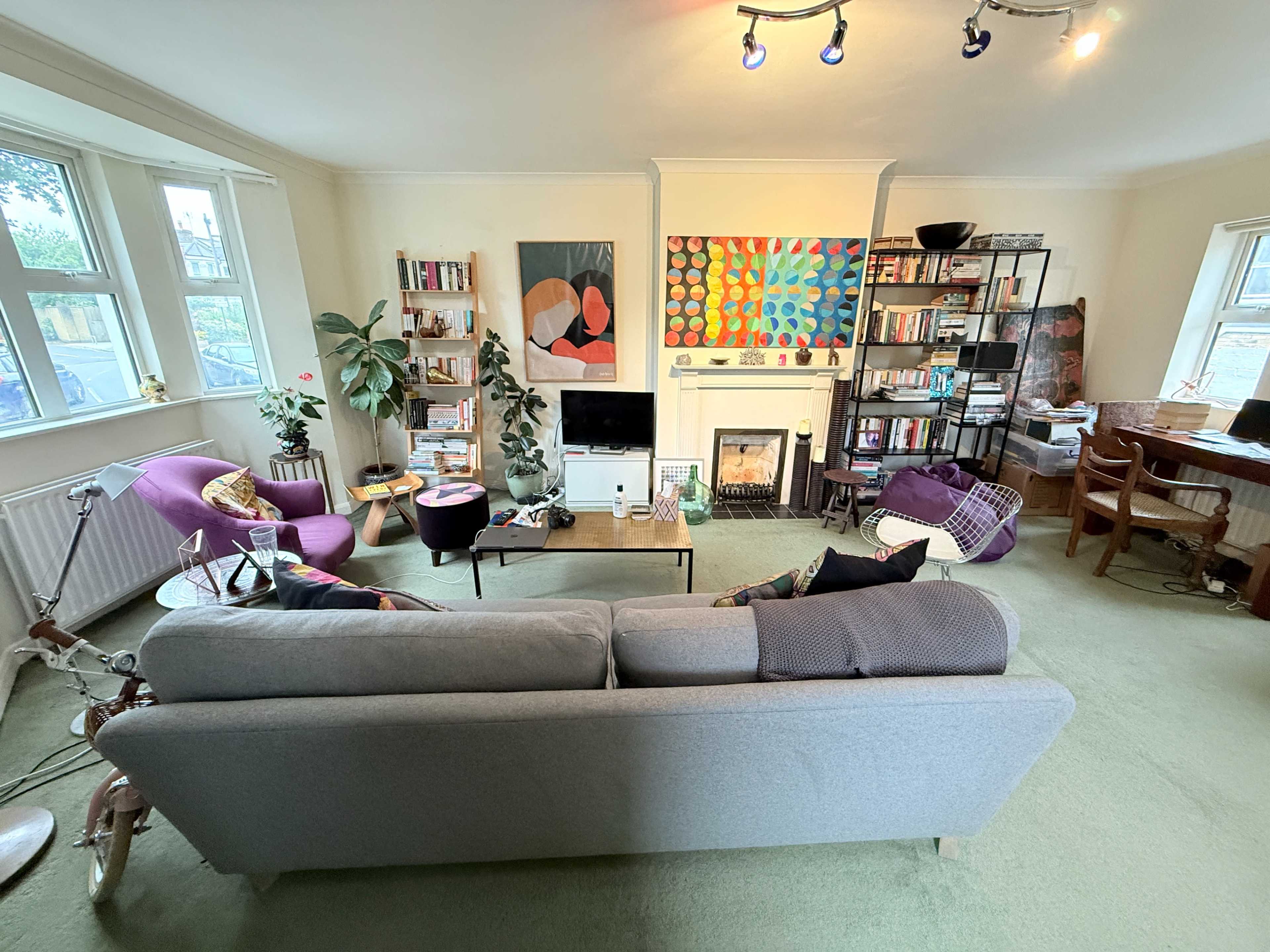 The image shows a cozy living room with a gray sofa, two purple armchairs, a television, shelves filled with books, and colorful artwork on the walls.