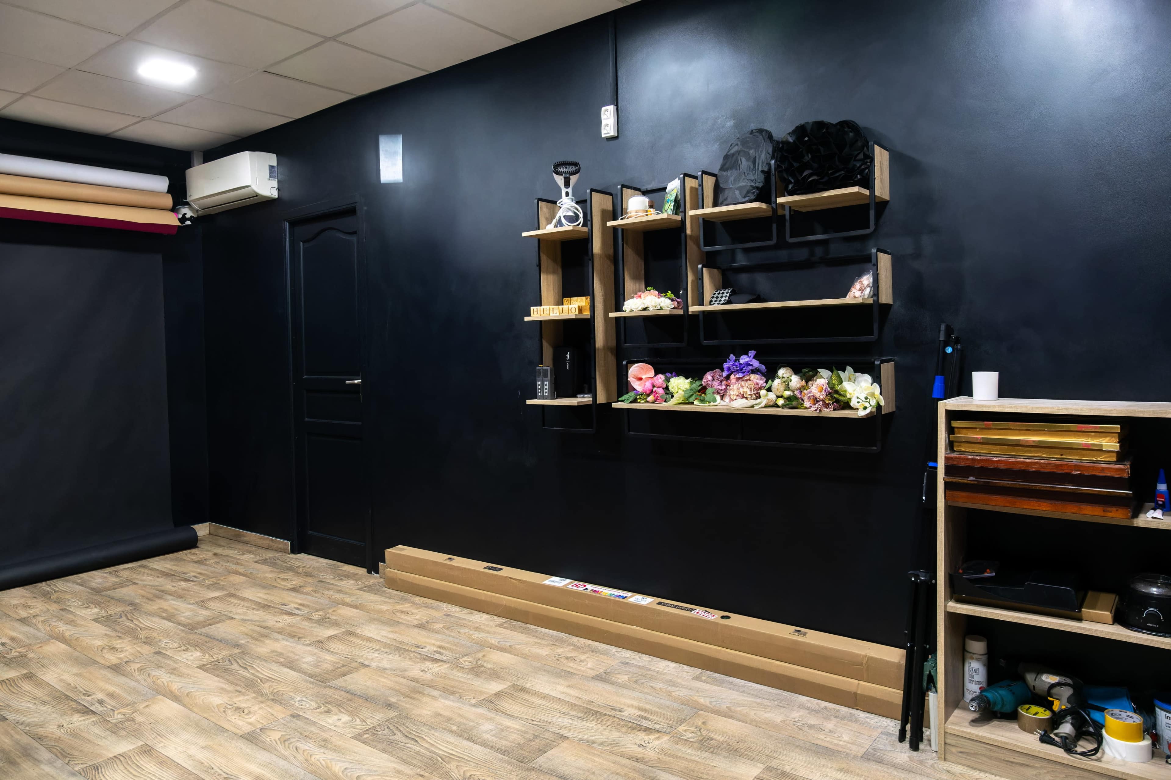 The image shows a dimly lit room with a dark wall, wooden shelves mounted on one side displaying decorative items and flowers, and a wooden floor with rolls of paper along the bottom.