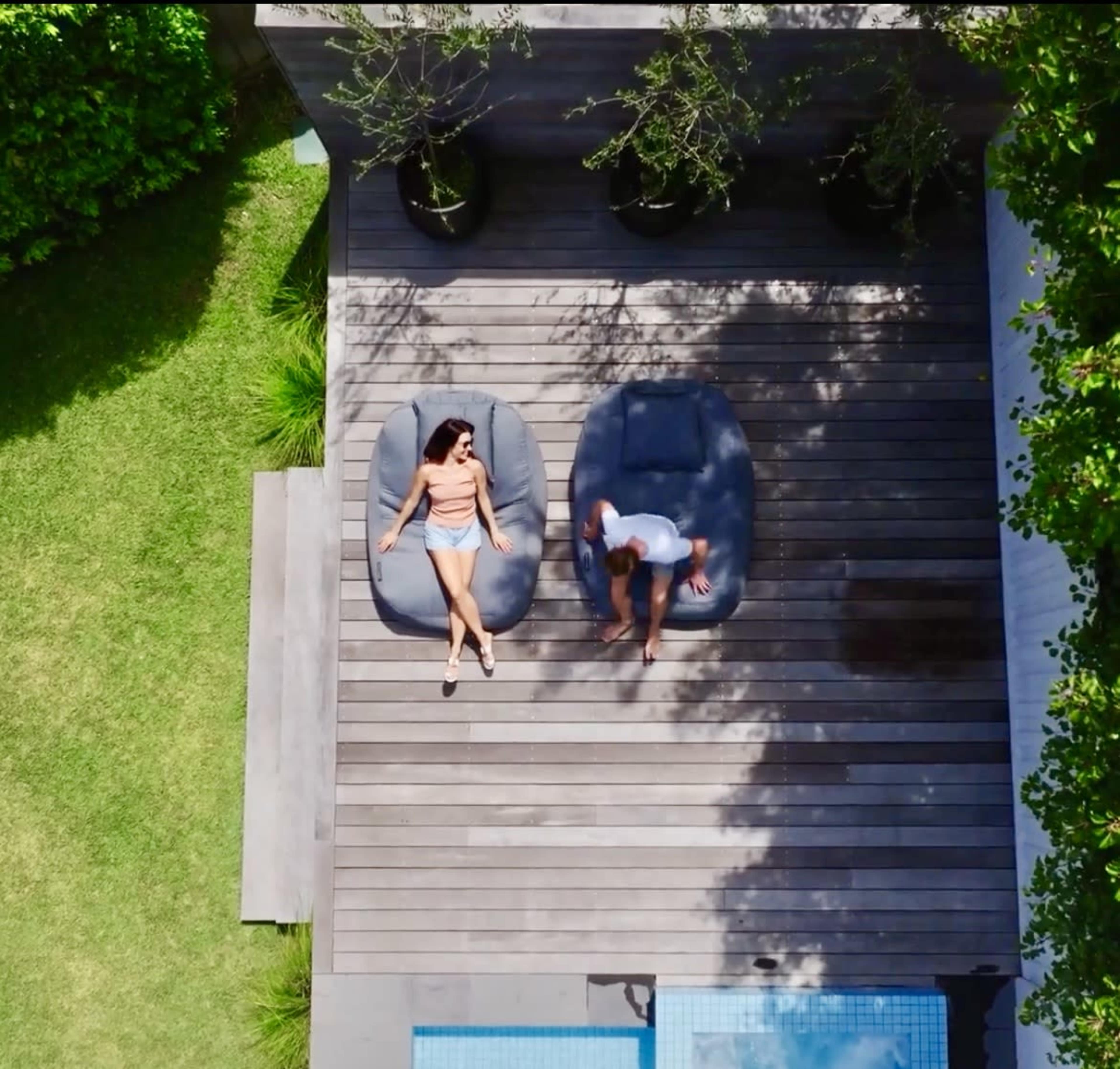 Two people relax on bean bag chairs beside a swimming pool on a wooden deck surrounded by greenery.