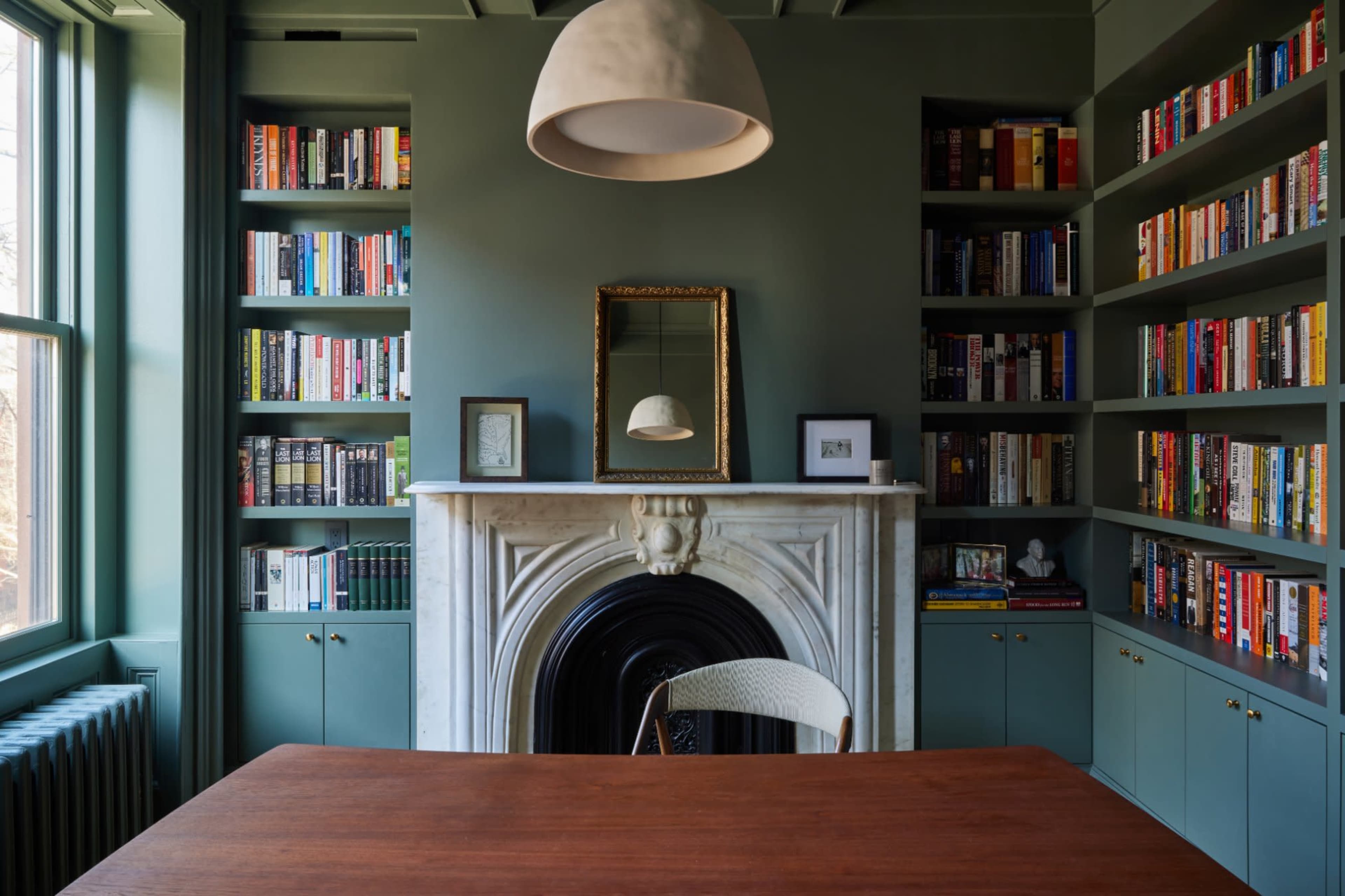 A cozy reading room features a wooden table, surrounded by bookshelves filled with books, and a decorative fireplace with a mirror above it.