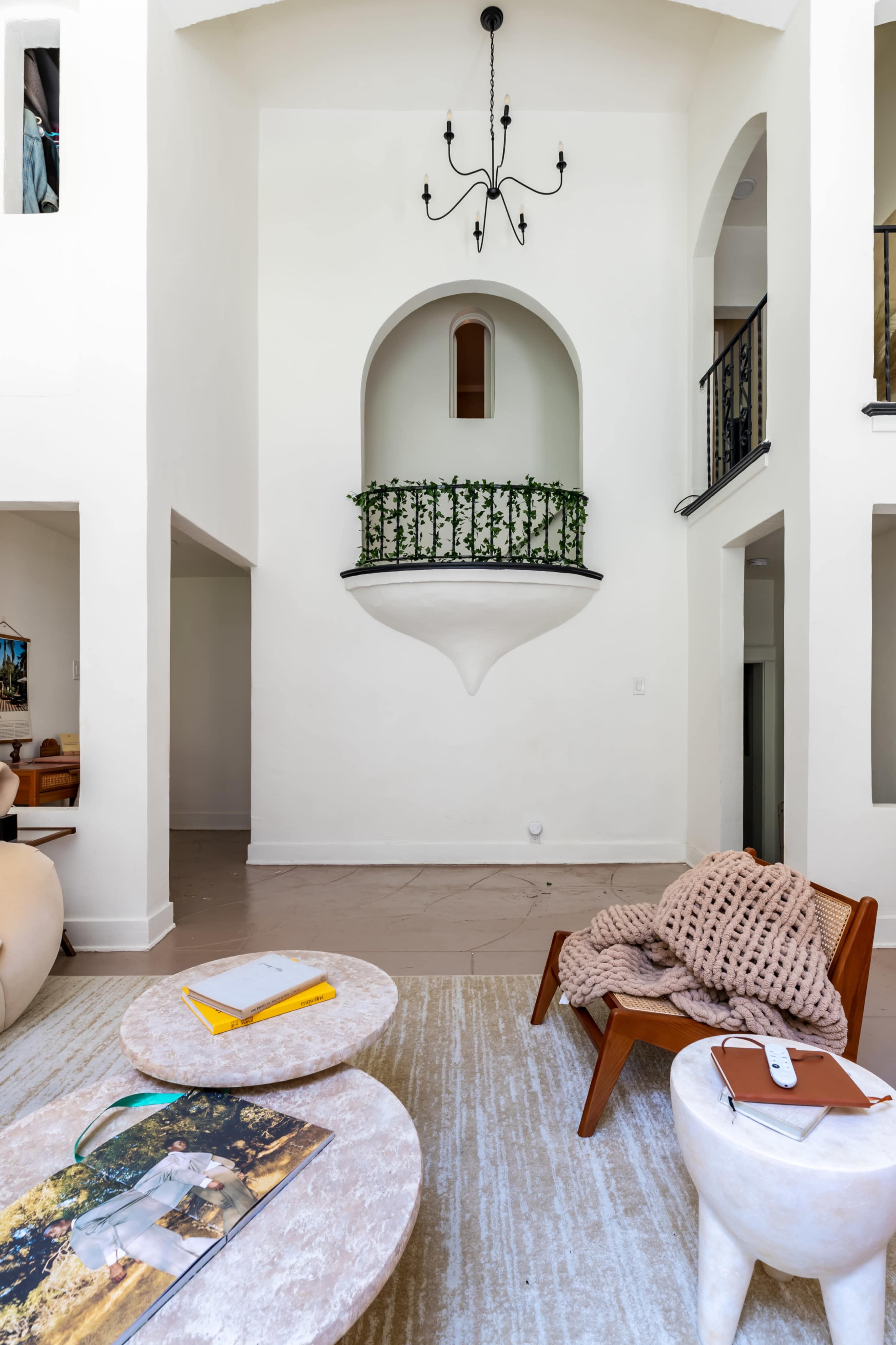A spacious living area with a high ceiling, featuring a balcony with greenery, a pair of round coffee tables, and a wooden chair draped with a blanket.