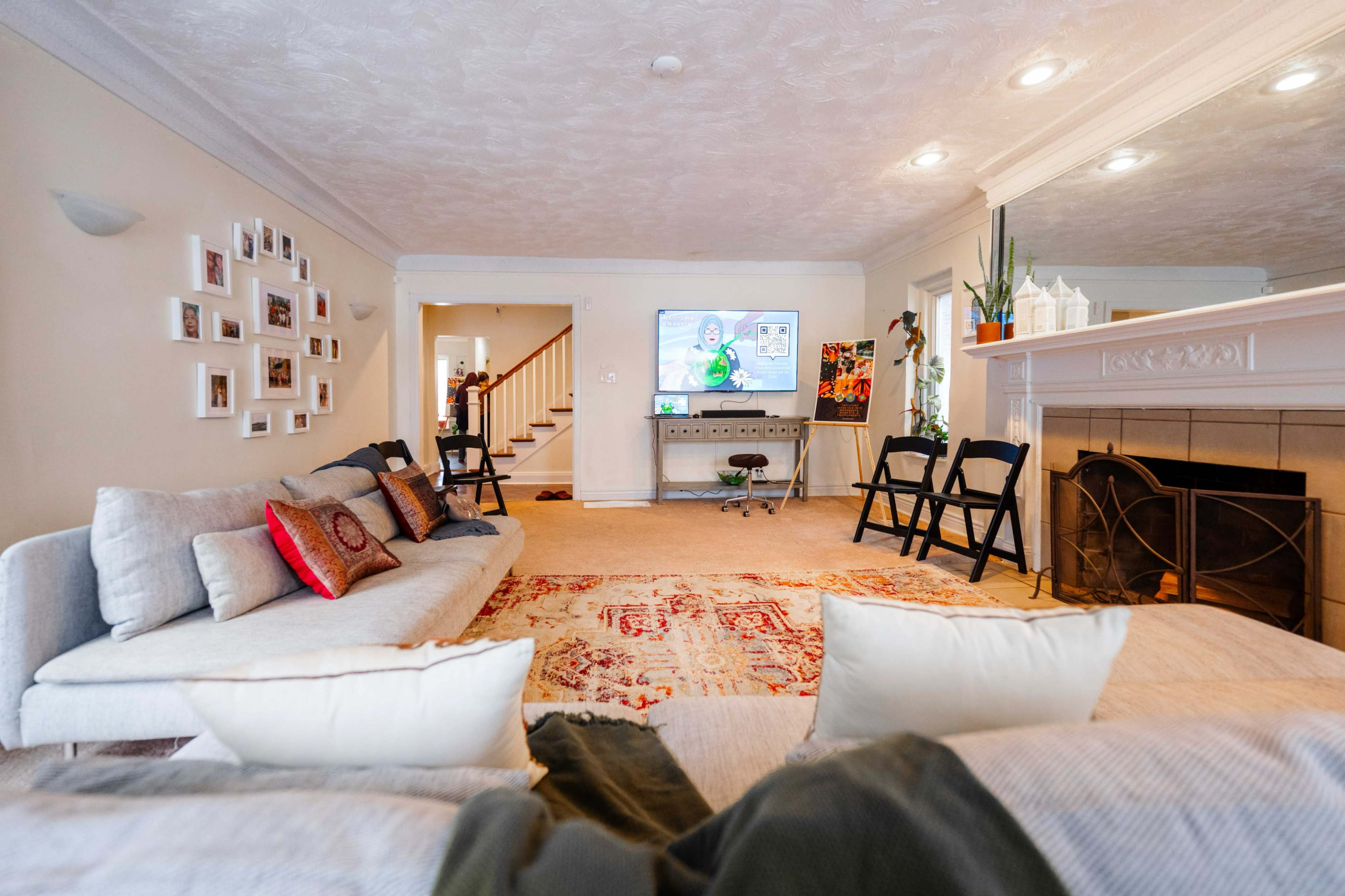 The living room features a large sectional sofa, a decorative area rug, a fireplace, and a television displaying animated content, with stairs leading to another level in the background.