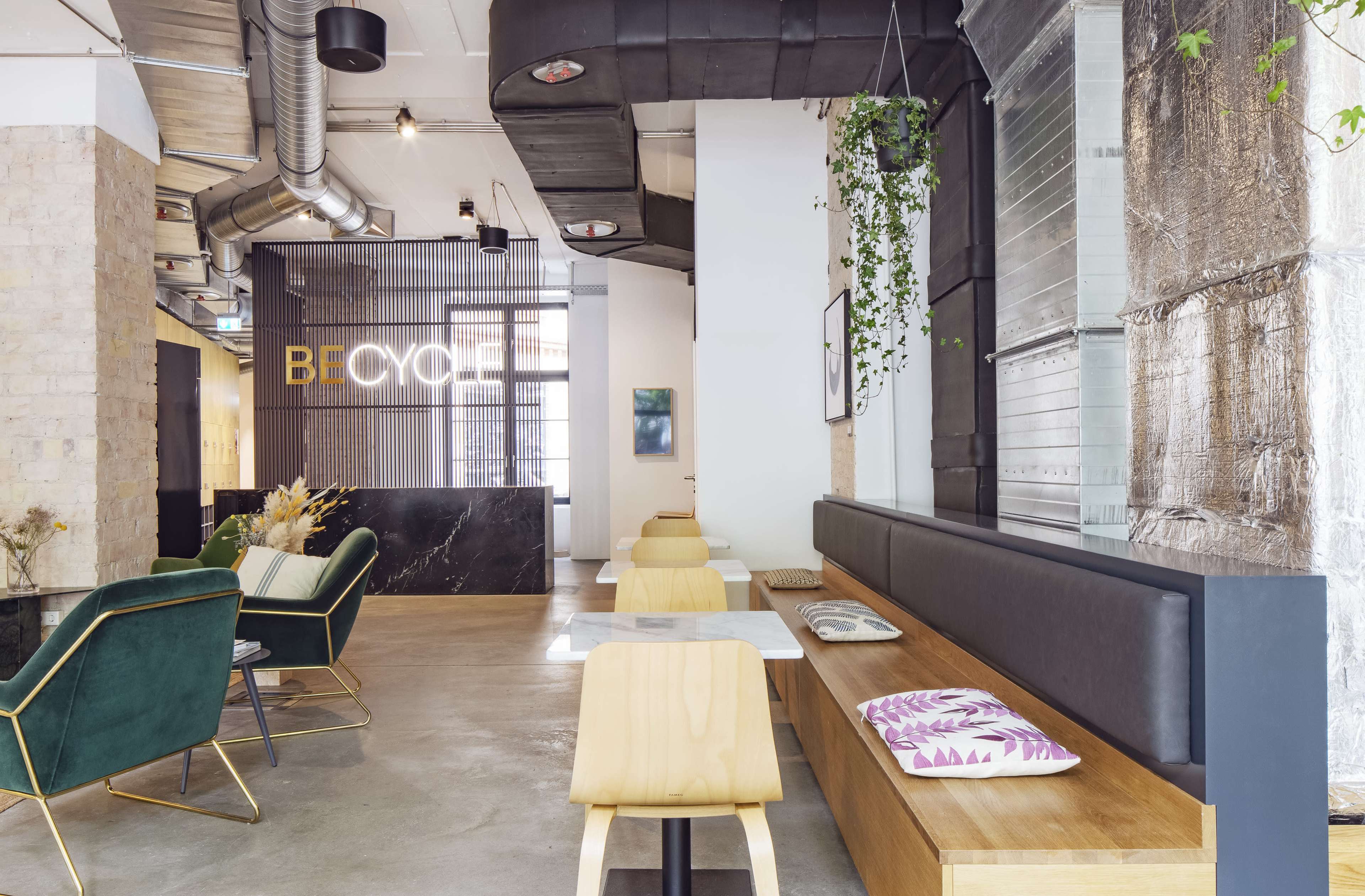 A modern interior space featuring a seating area with wooden tables and green chairs, along with a large sign that reads "BECYCLE" on the wall.