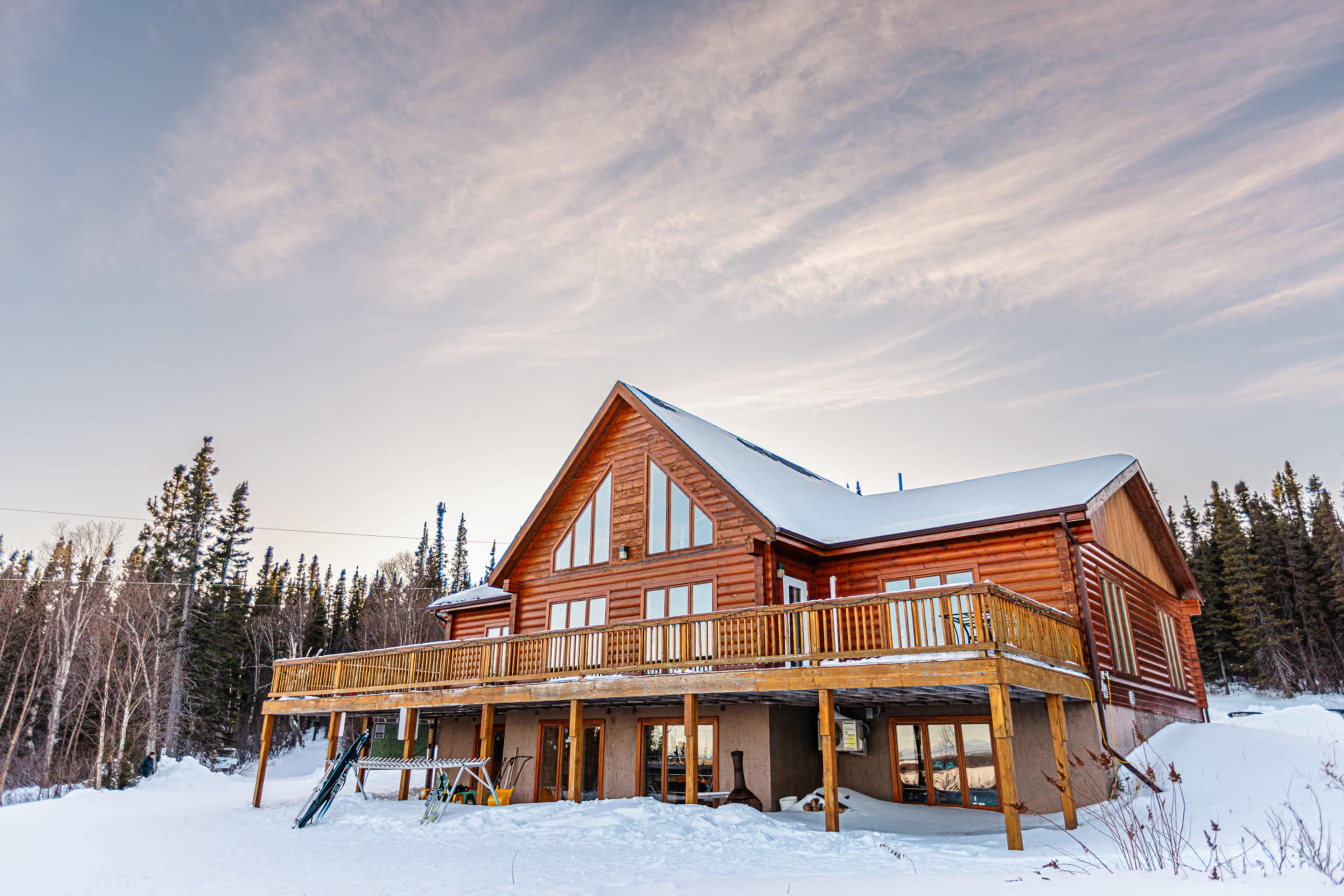 A large wooden cabin with a wraparound deck and expansive windows is situated in a snowy landscape surrounded by trees.