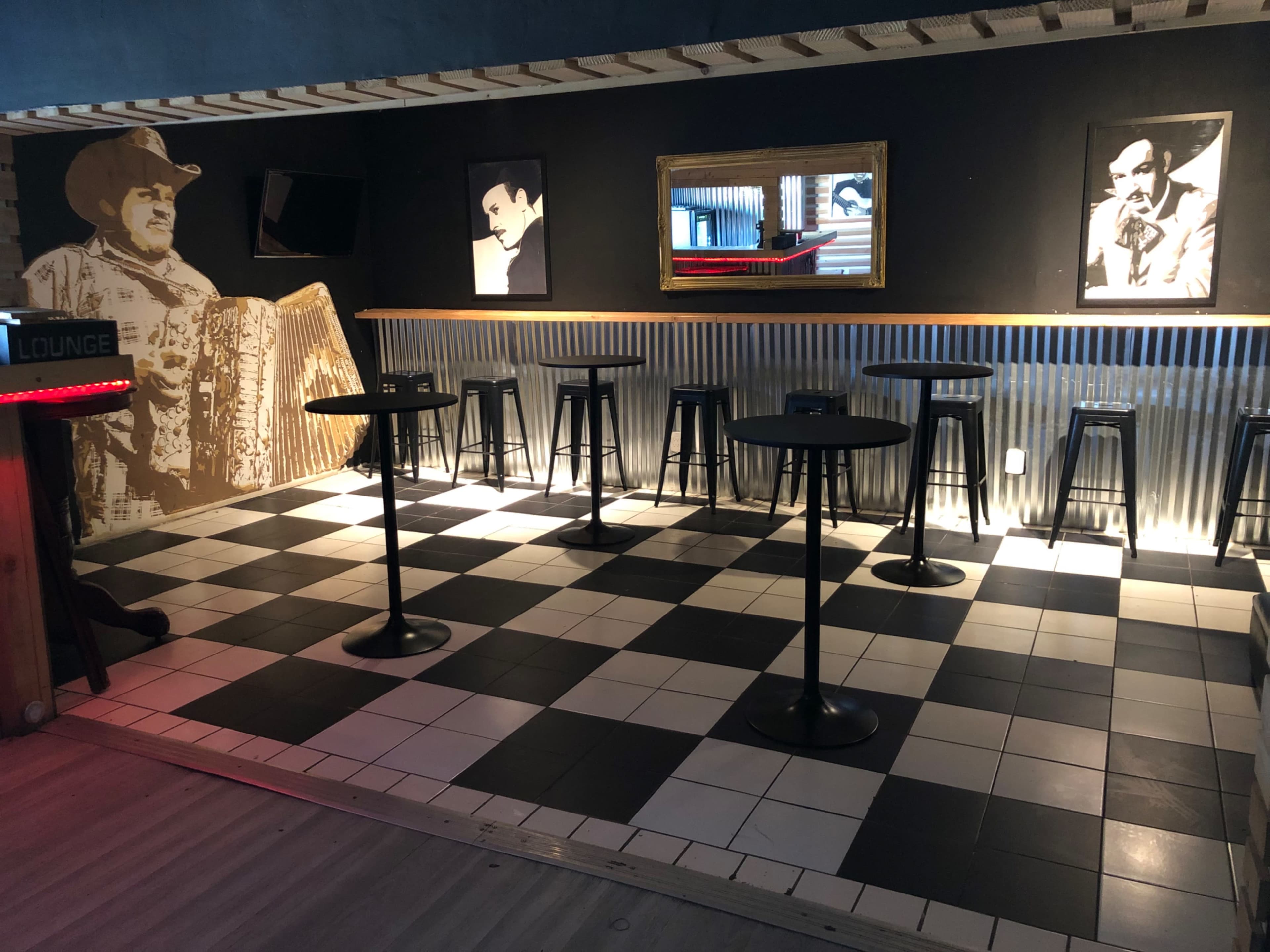 A bar lounge with a checkered black and white floor, several metal tables and stools, and a wall featuring a mural and framed portraits.