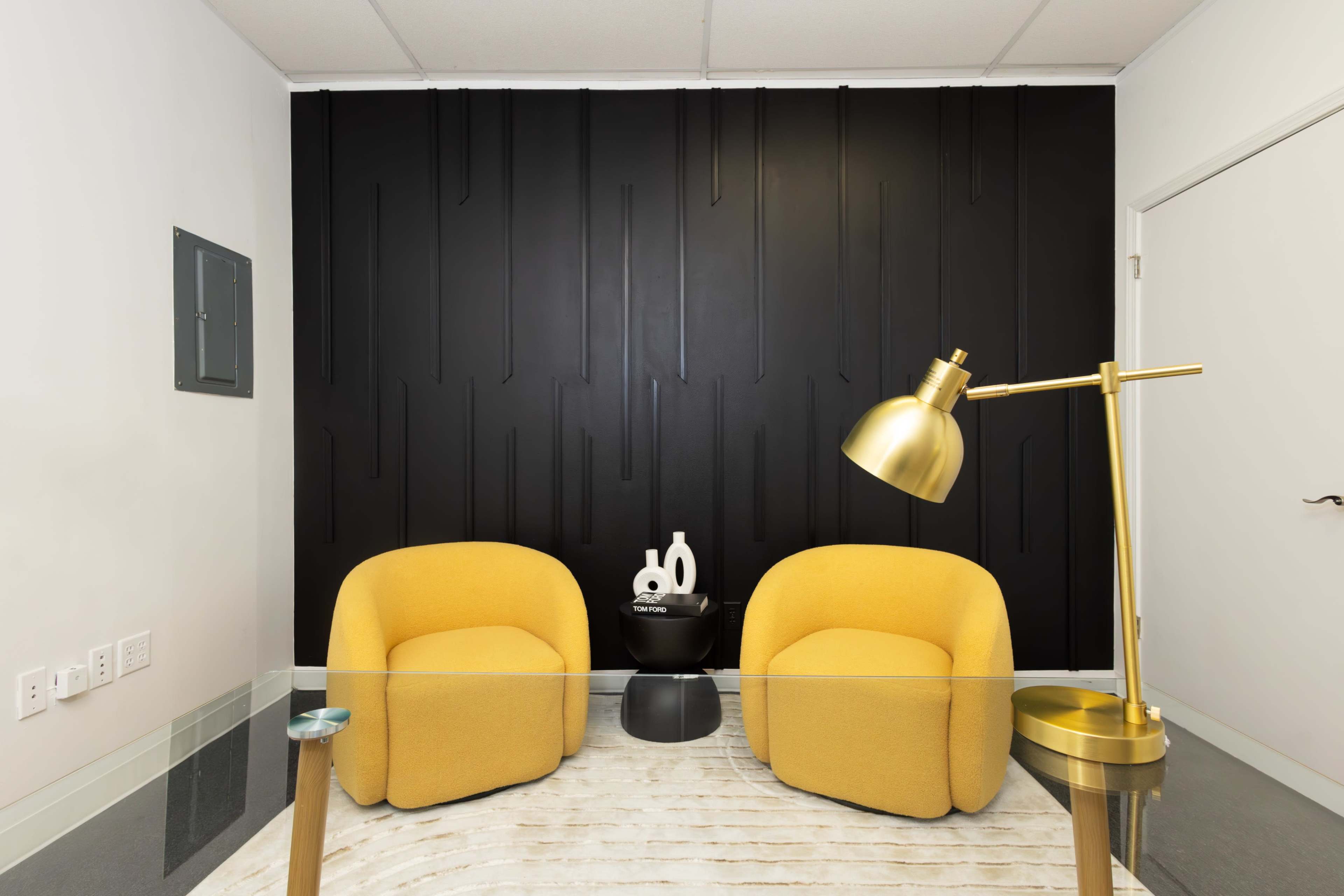The image shows a modern office space featuring two yellow armchairs, a glass table, a golden lamp, and a textured black wall.
