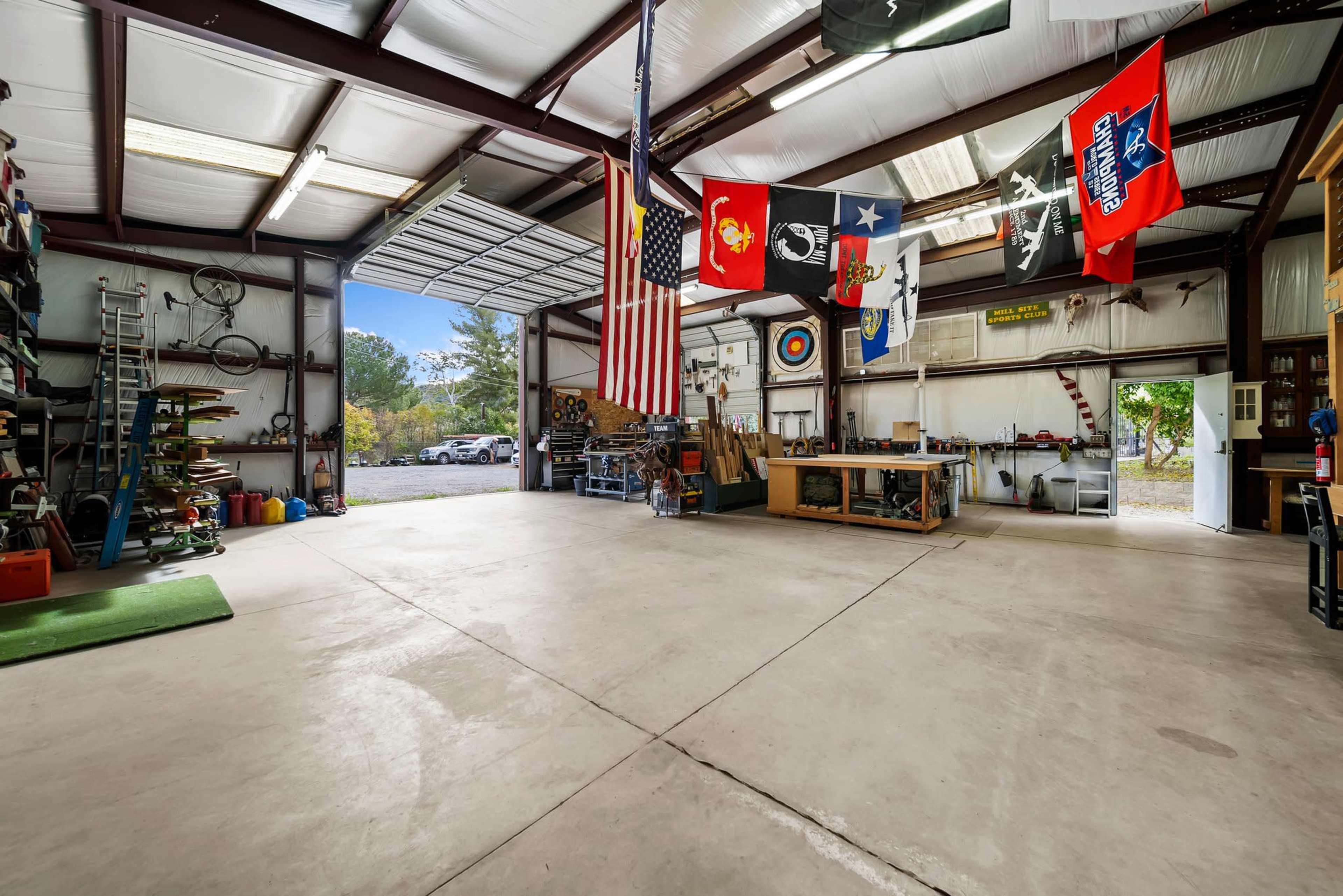 The image shows an expansive industrial garage or workshop with metal framing, concrete flooring, and various tools and equipment arranged along the walls, along with multiple flags hanging from the ceiling.