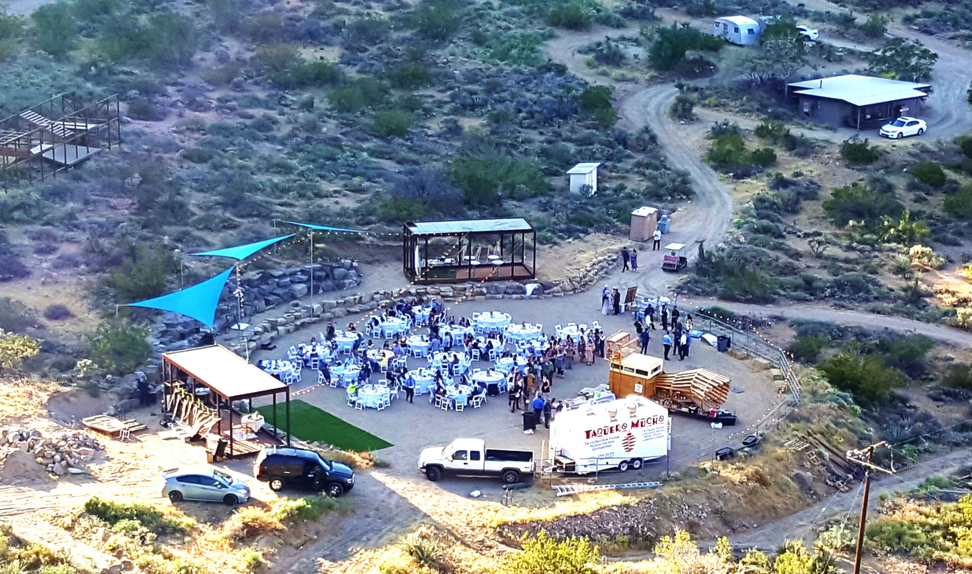 An outdoor gathering is set up in a cleared area with tables and chairs, surrounded by desert vegetation and structures in the background.