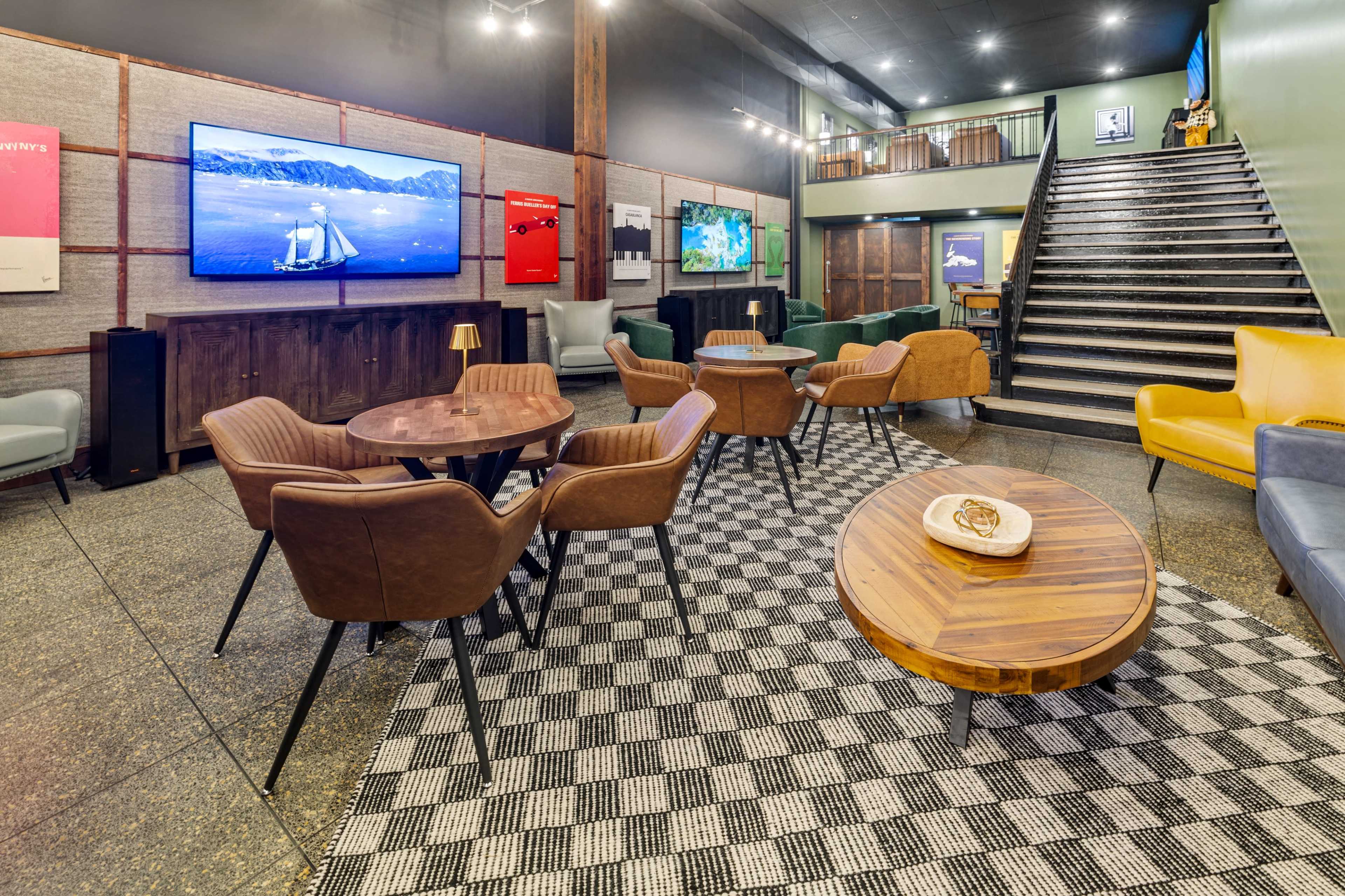 The image shows a modern lounge area with several seating arrangements, including wooden tables and upholstered chairs, along with a large TV mounted on the wall.
