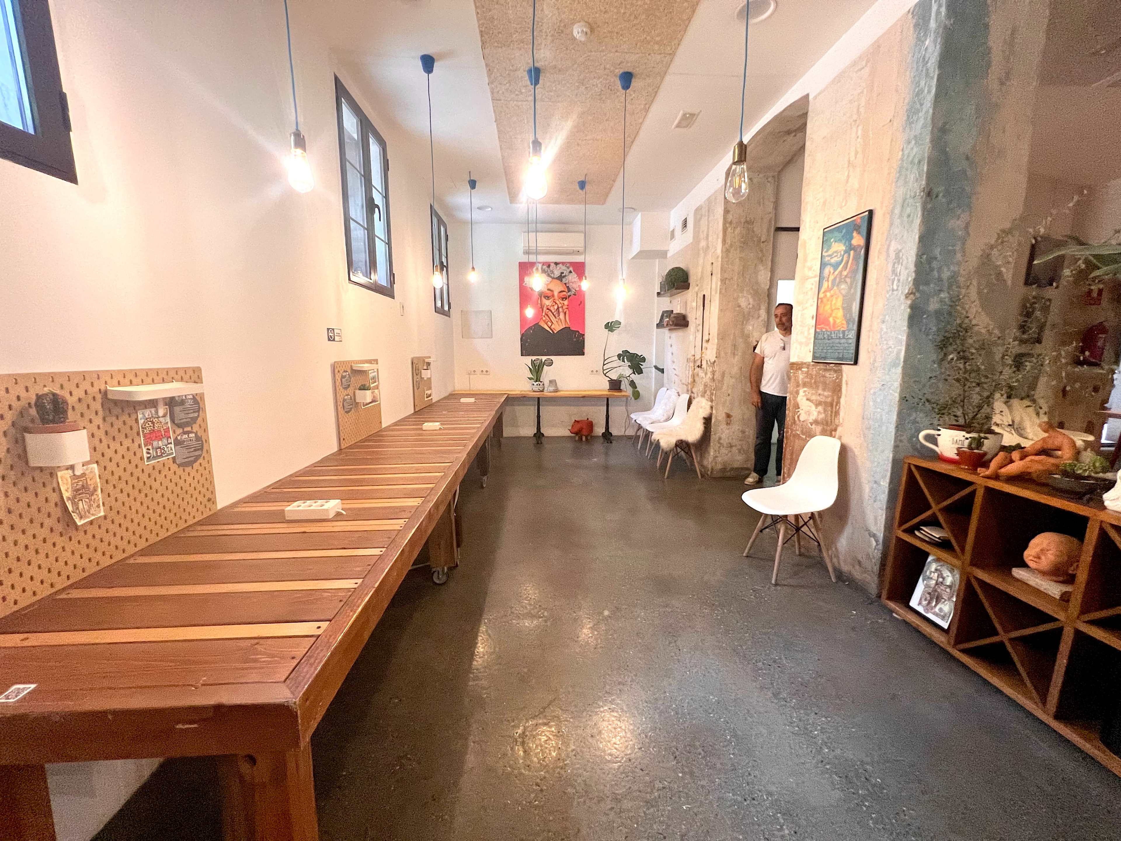The image shows a modern interior space with a long wooden table, minimalist white chairs, and exposed light bulbs hanging from the ceiling.