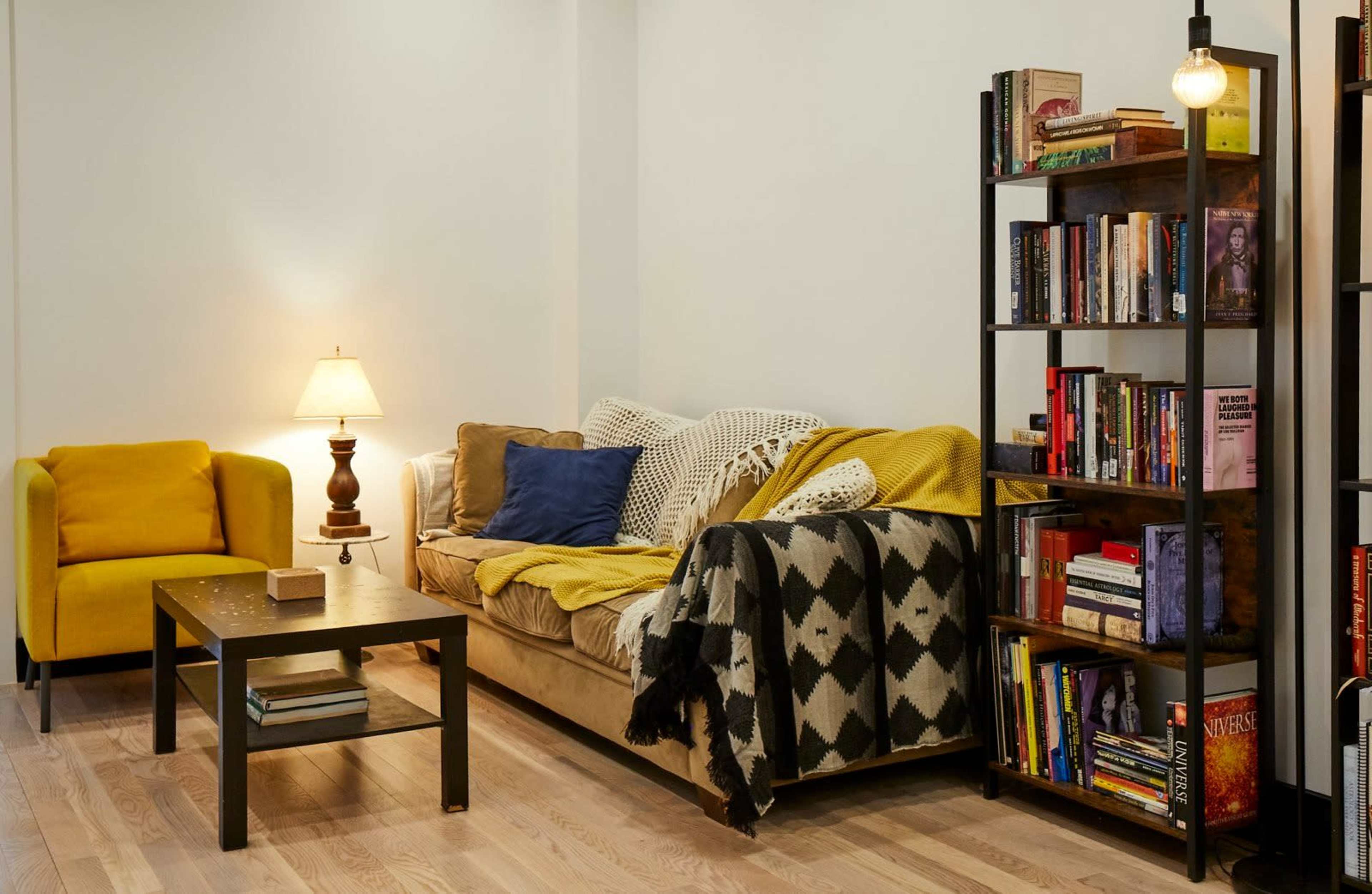 A cozy living room with a yellow armchair, a beige sofa covered in blankets, a small table, and a bookshelf filled with books.