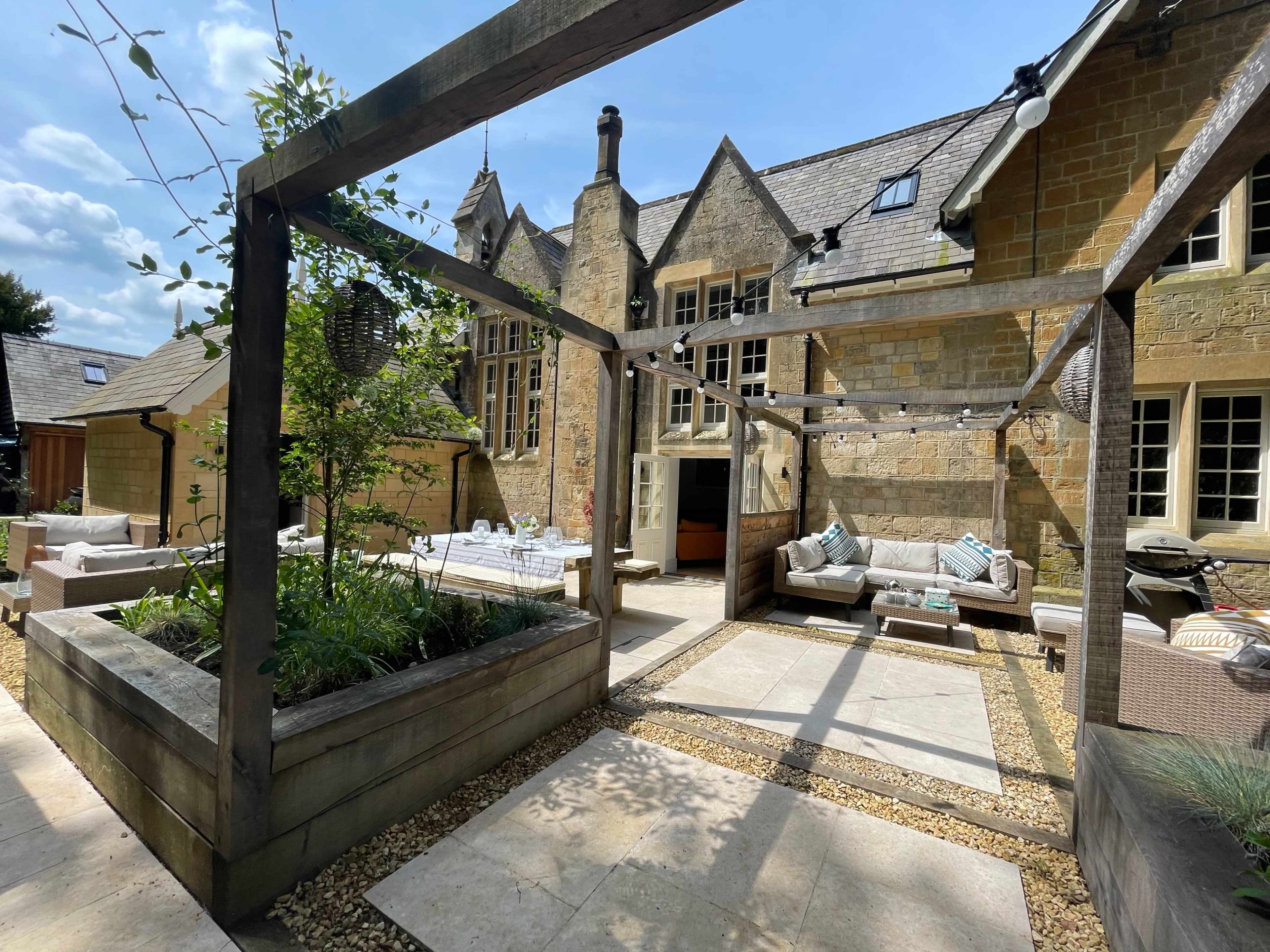 The image shows a landscaped courtyard with stone walls, wooden pergolas, and seating areas, set against a backdrop of a large stone building under a clear blue sky.