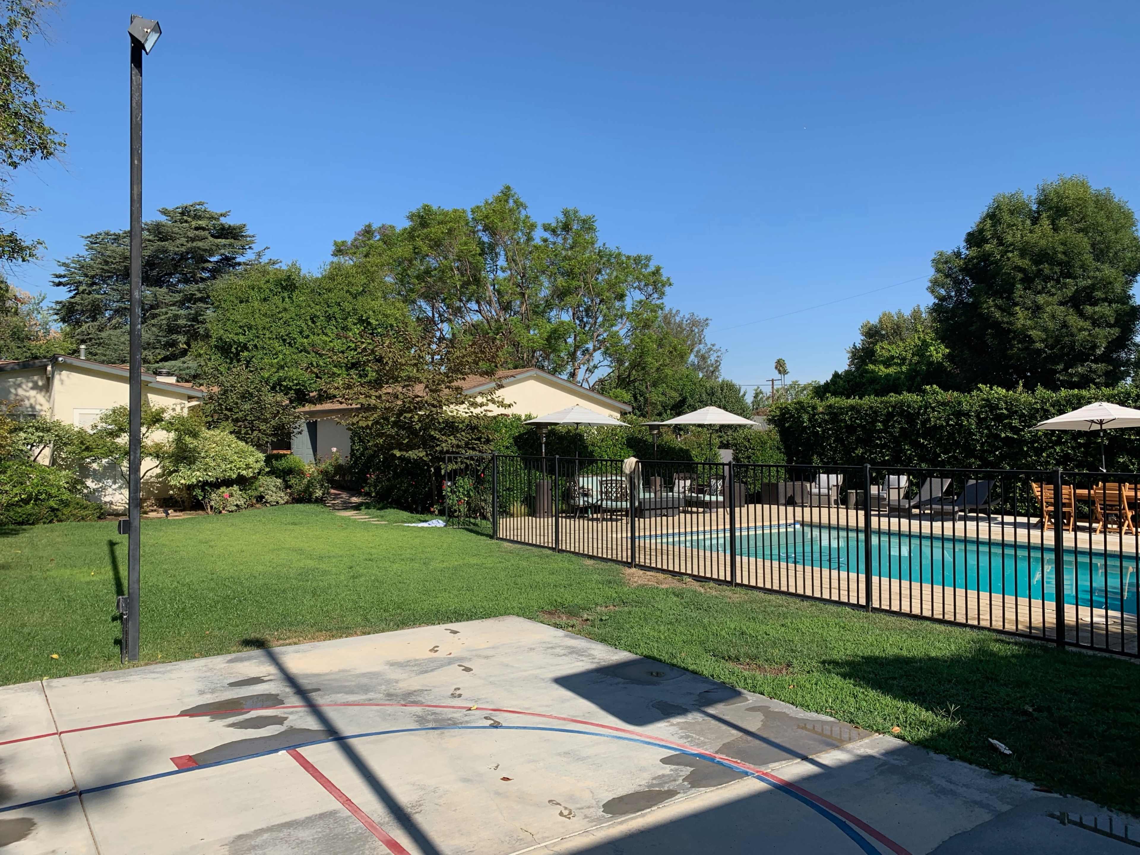 A backyard with a basketball hoop, grassy area, and a swimming pool surrounded by a fence and lounge chairs.