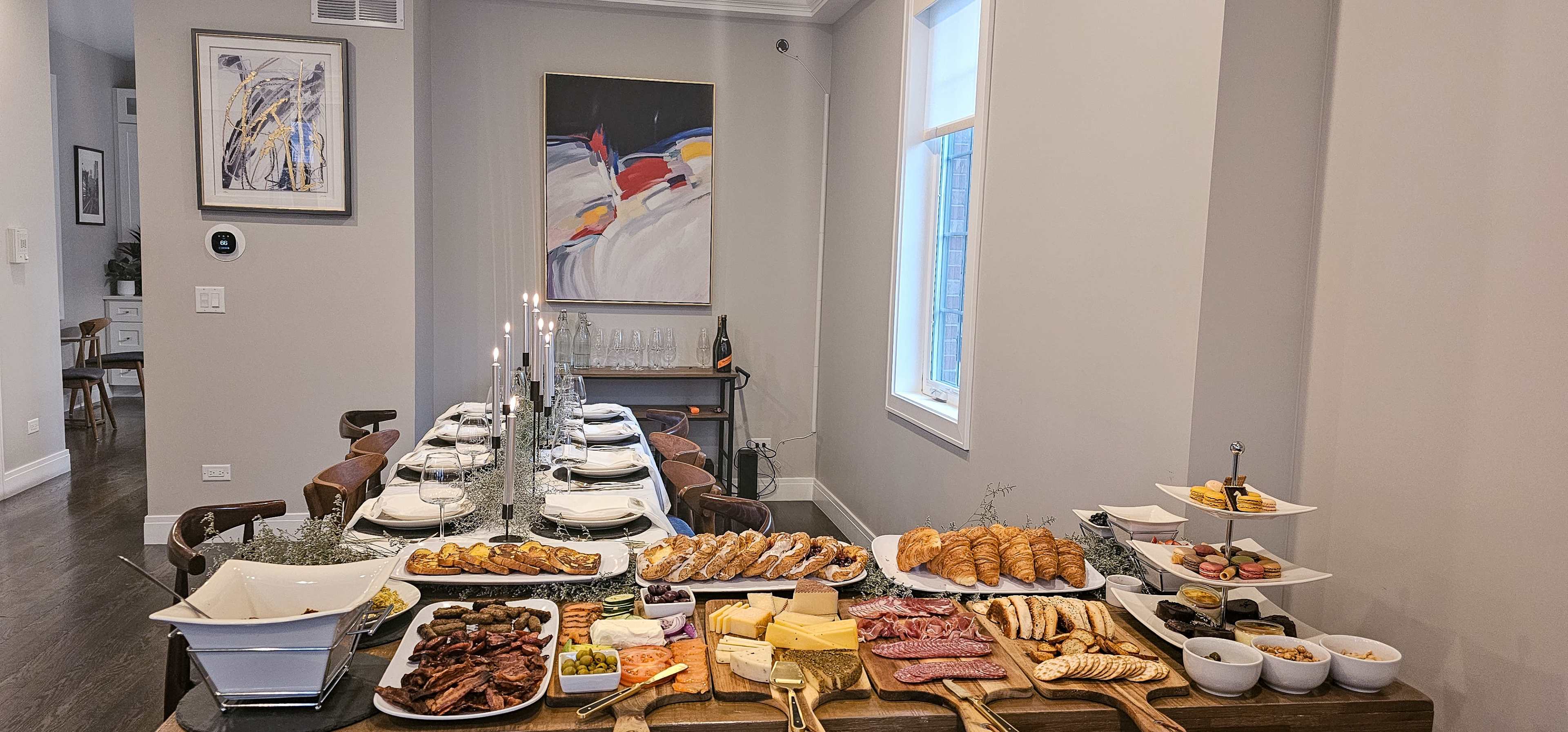 A long table is set with a variety of food, including cheeses, meats, pastries, and vegetables, alongside wine glasses and candles in a well-lit dining room.