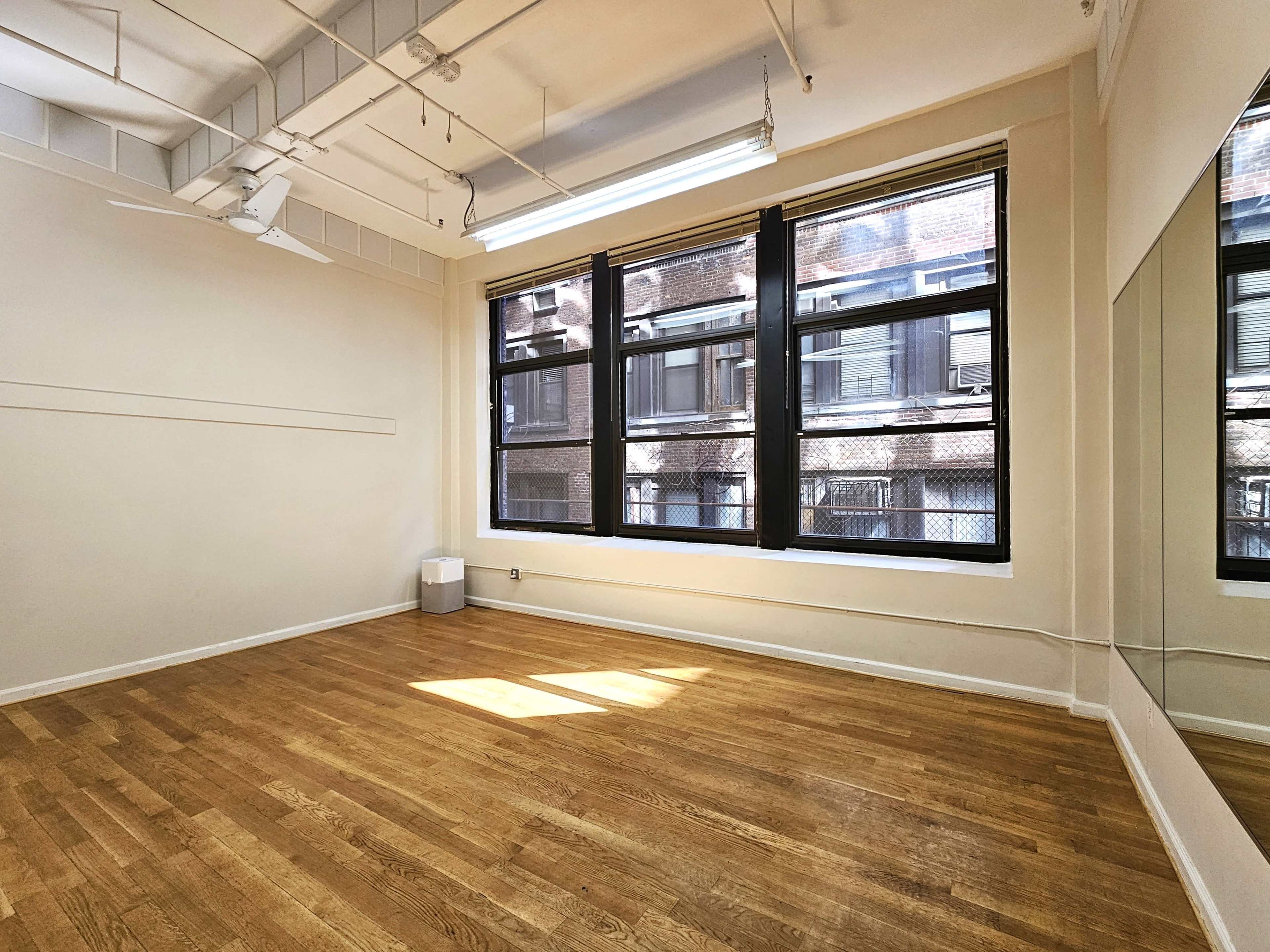 The image shows a spacious room with wooden floors, large windows, and a mirror on one wall.