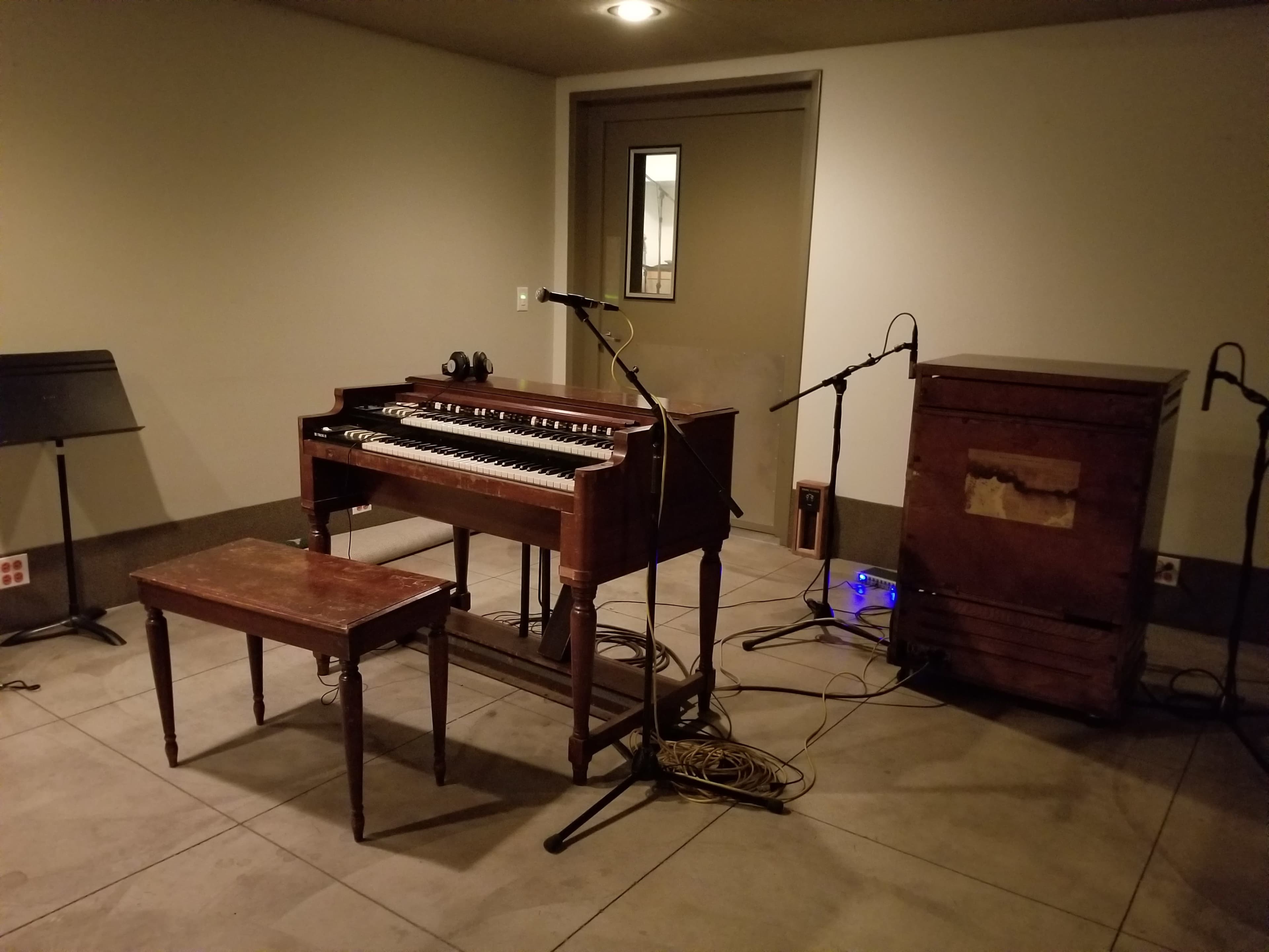 A vintage organ with a small wooden bench sits in an empty room next to a vintage amplifier and microphones on stands.