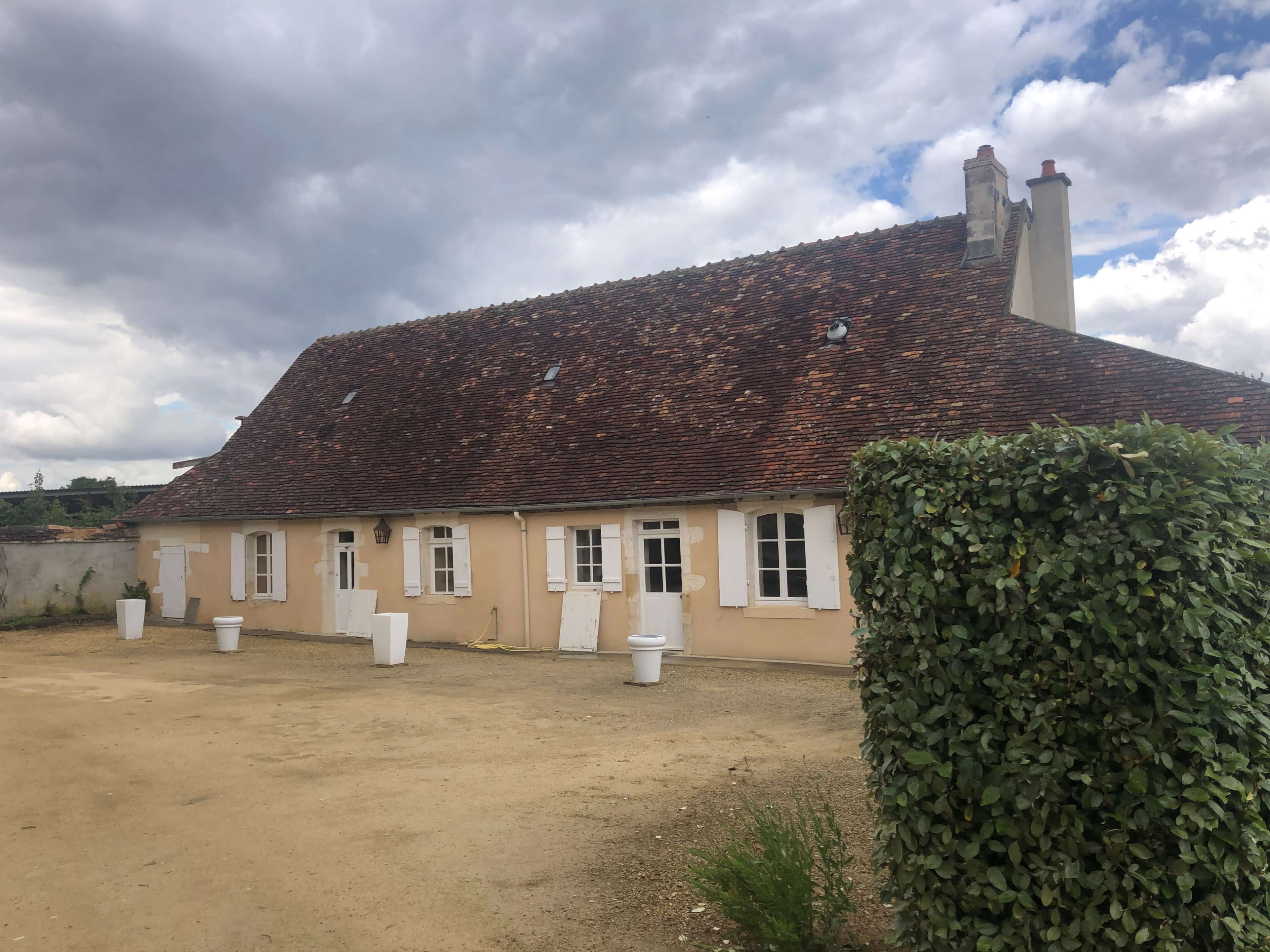 The image shows a single-story house with a sloped roof and several windows, surrounded by a gravel area and a trimmed hedge under an overcast sky.