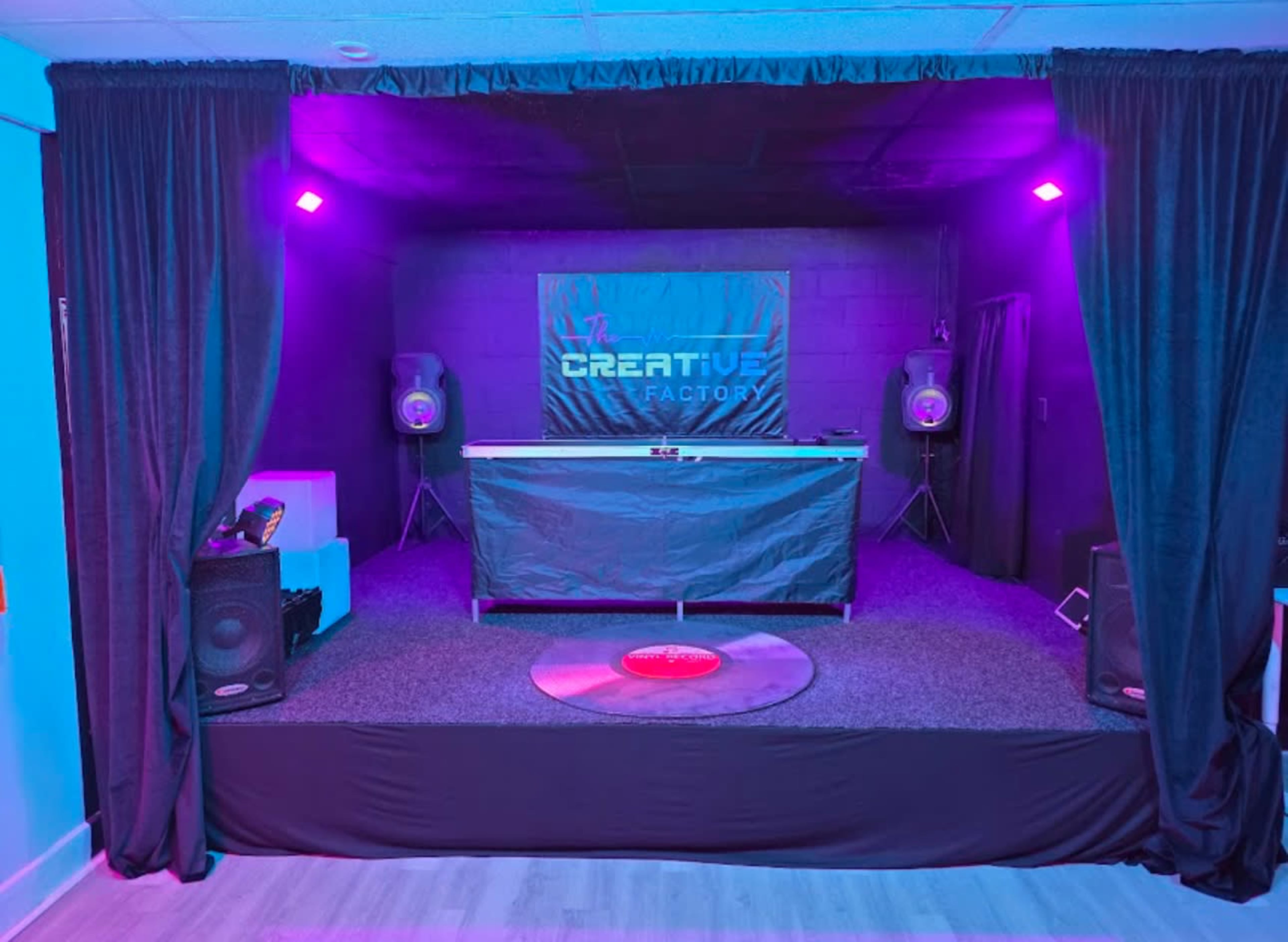 The image shows a dimly lit creative studio space with a stage featuring a black table, large speakers, and a backdrop with the text "CREATIVE FACTORY."