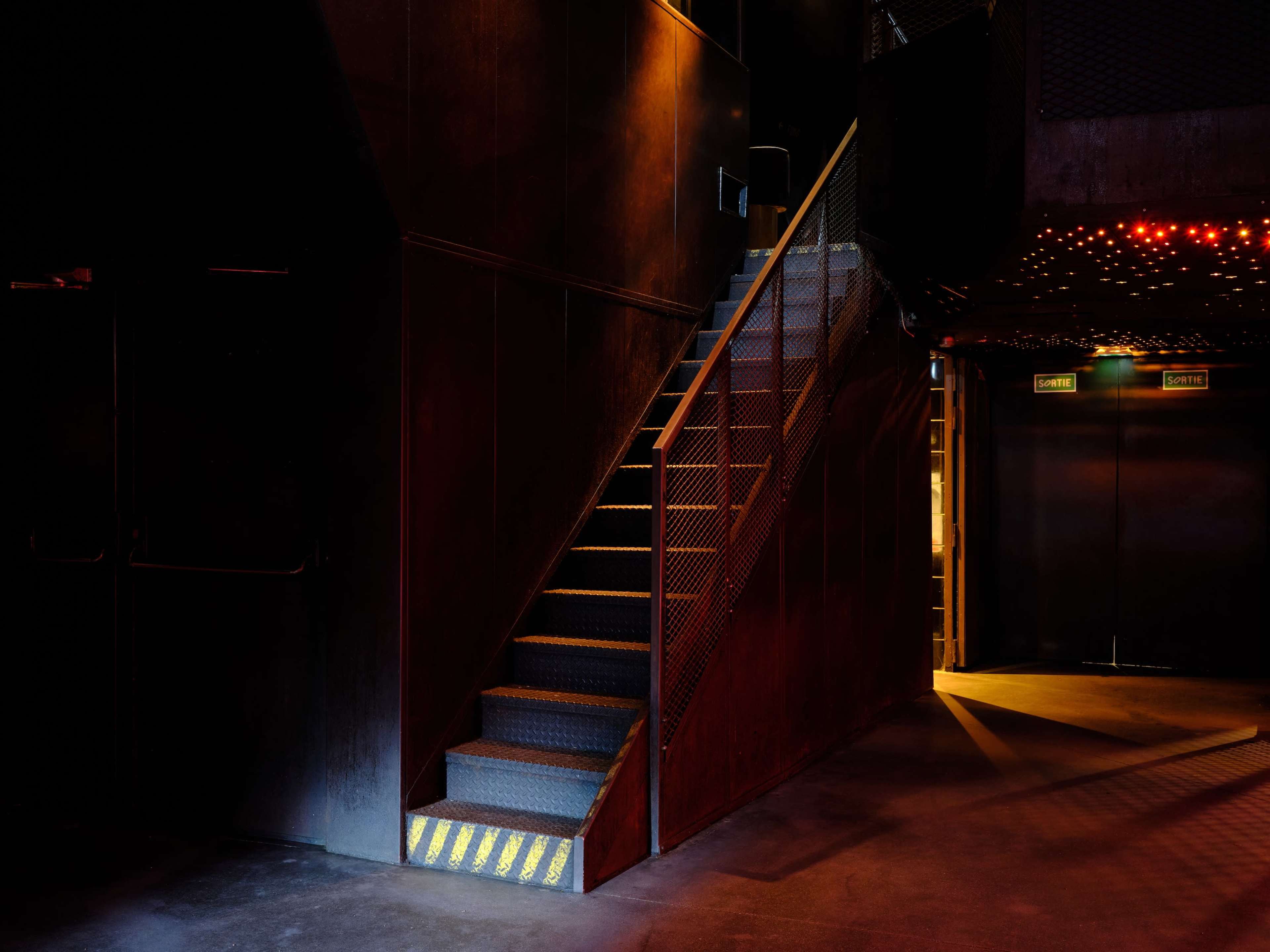 A metal staircase ascends in a dimly lit industrial space, with the glow of lights from the upper level illuminating the surroundings.