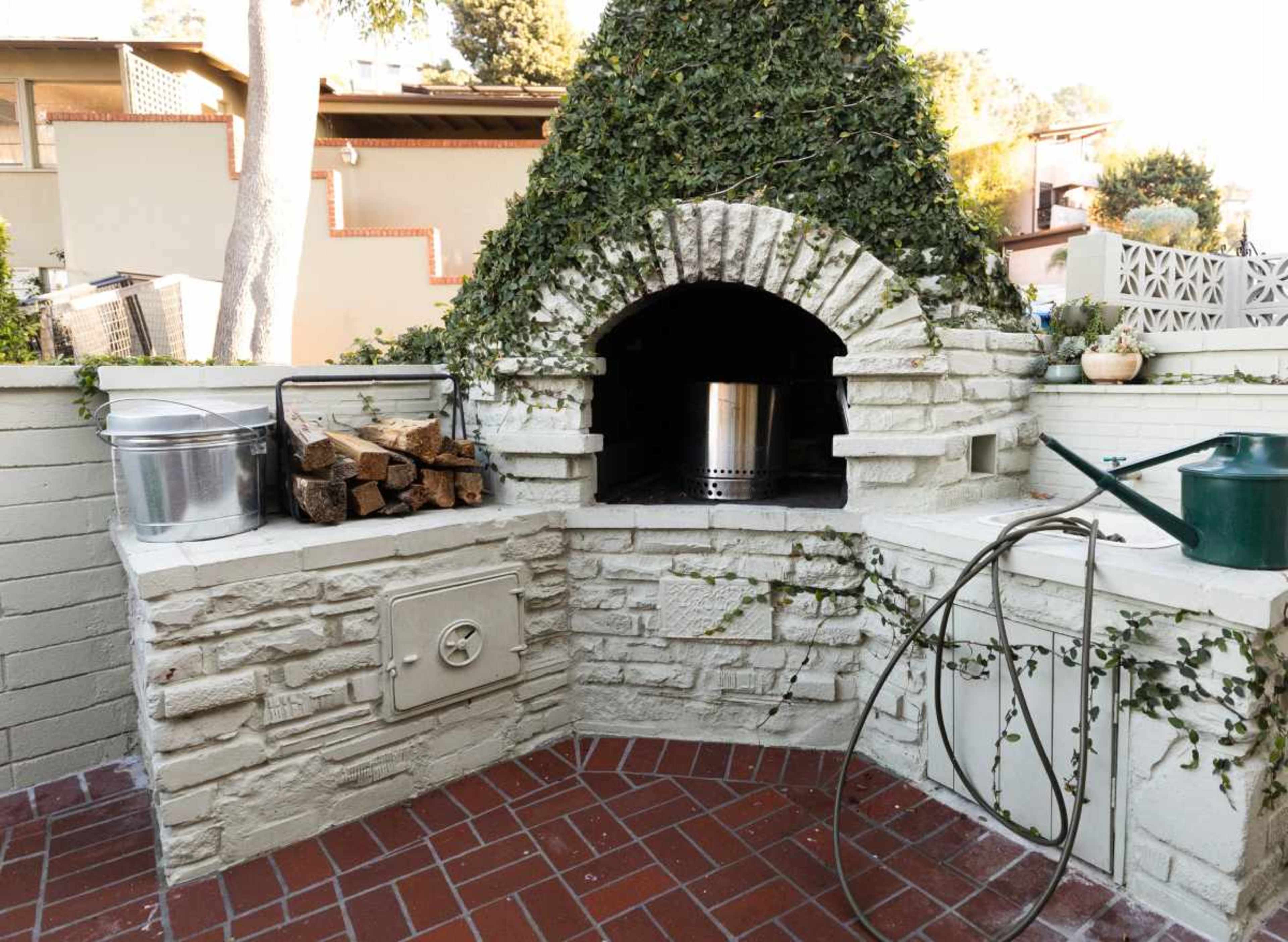 The image shows an outdoor stone pizza oven adorned with ivy, surrounded by a stack of firewood, a metal bucket, a watering can, and a garden hose.