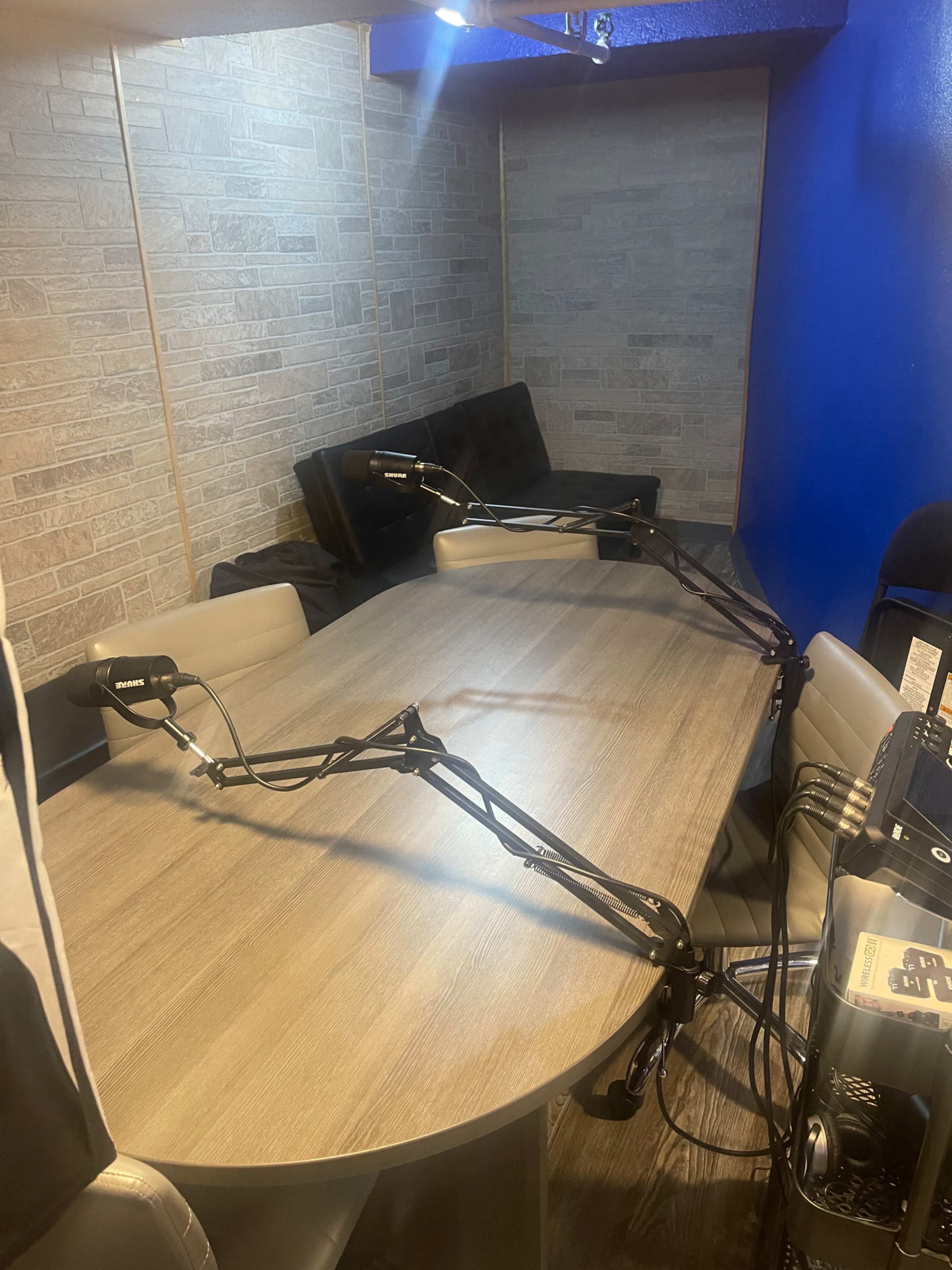 A long wooden table with several chairs is set up in a room, featuring two adjustable microphones mounted on arms, against a backdrop of a textured wall.