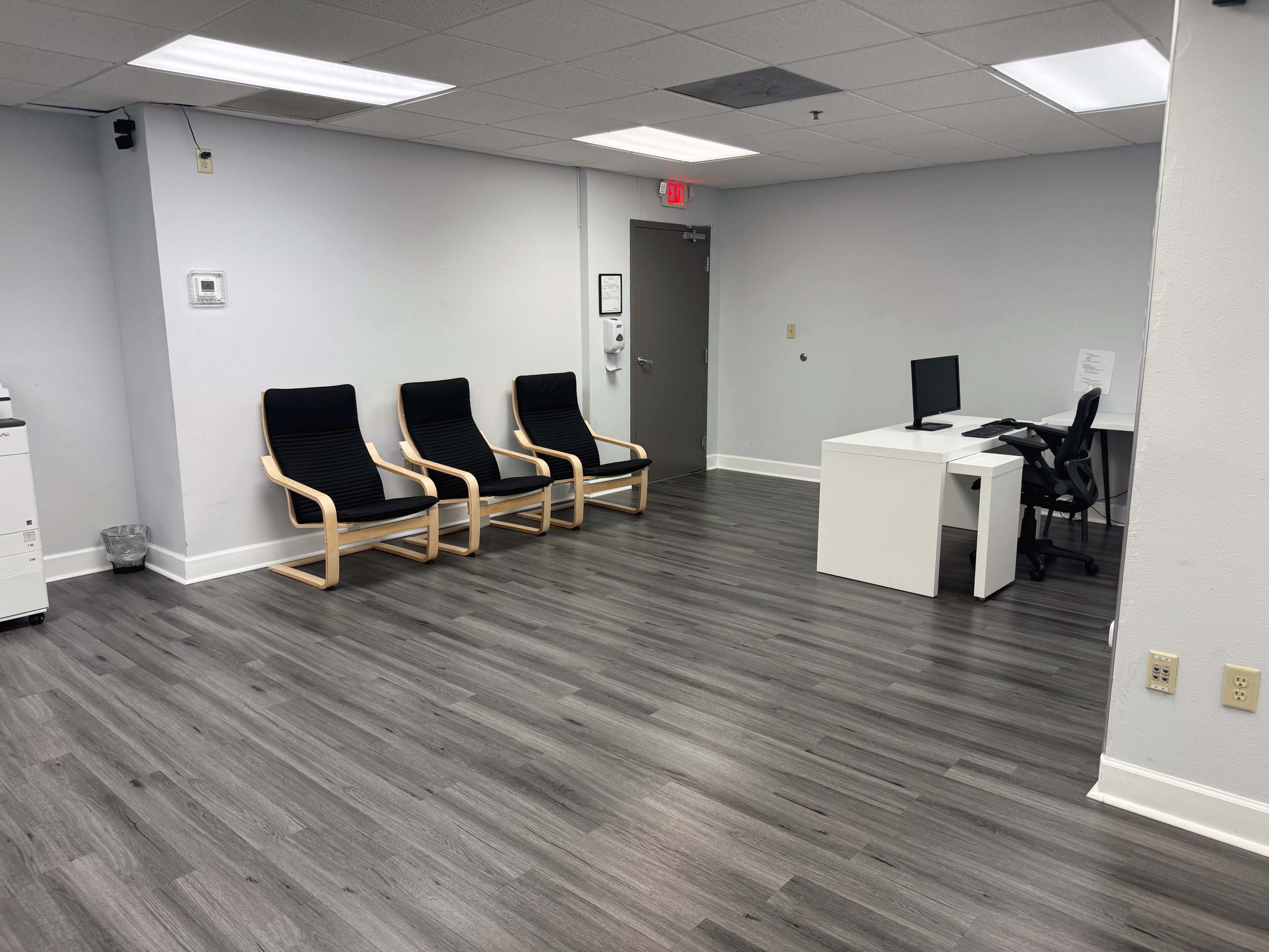 The image shows a modern waiting room with three chairs, a small desk with a computer, and a printer in the corner.