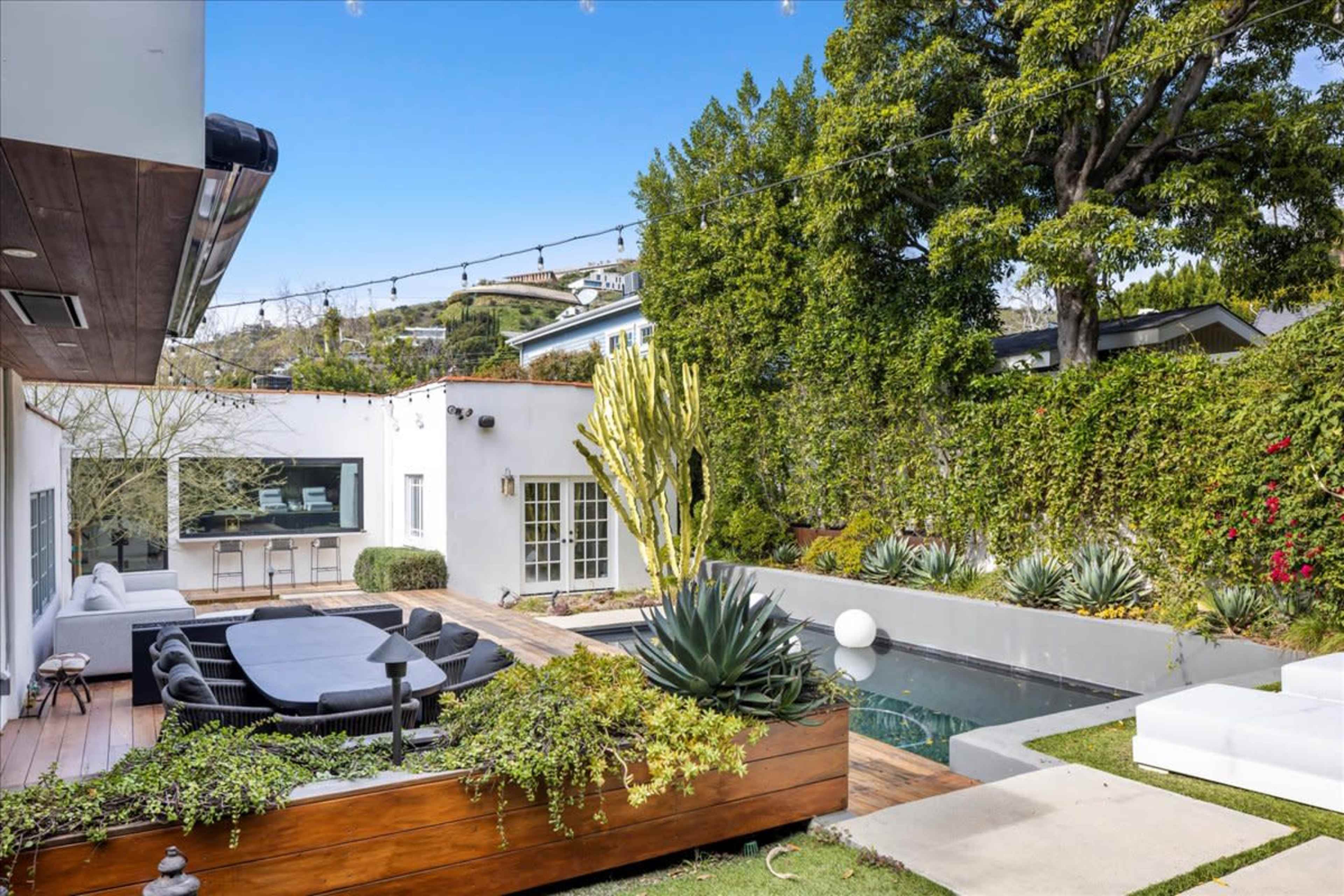 The image shows a modern outdoor living space with a swimming pool, seating area, and various plants surrounded by greenery.