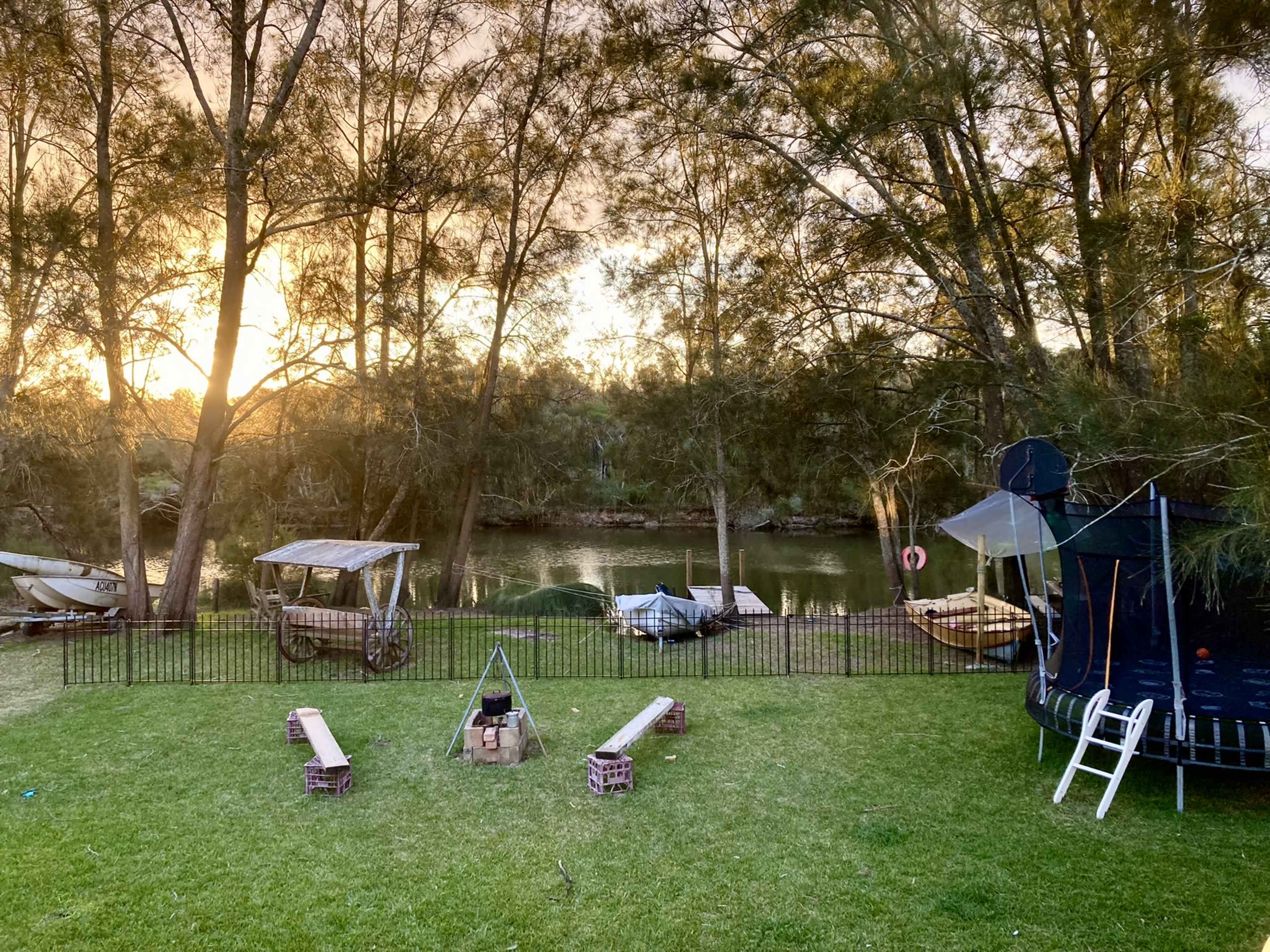 The scene features a grassy area by a river with a trampoline, wooden benches arranged around a fire pit, and several boats moored nearby under tall trees.
