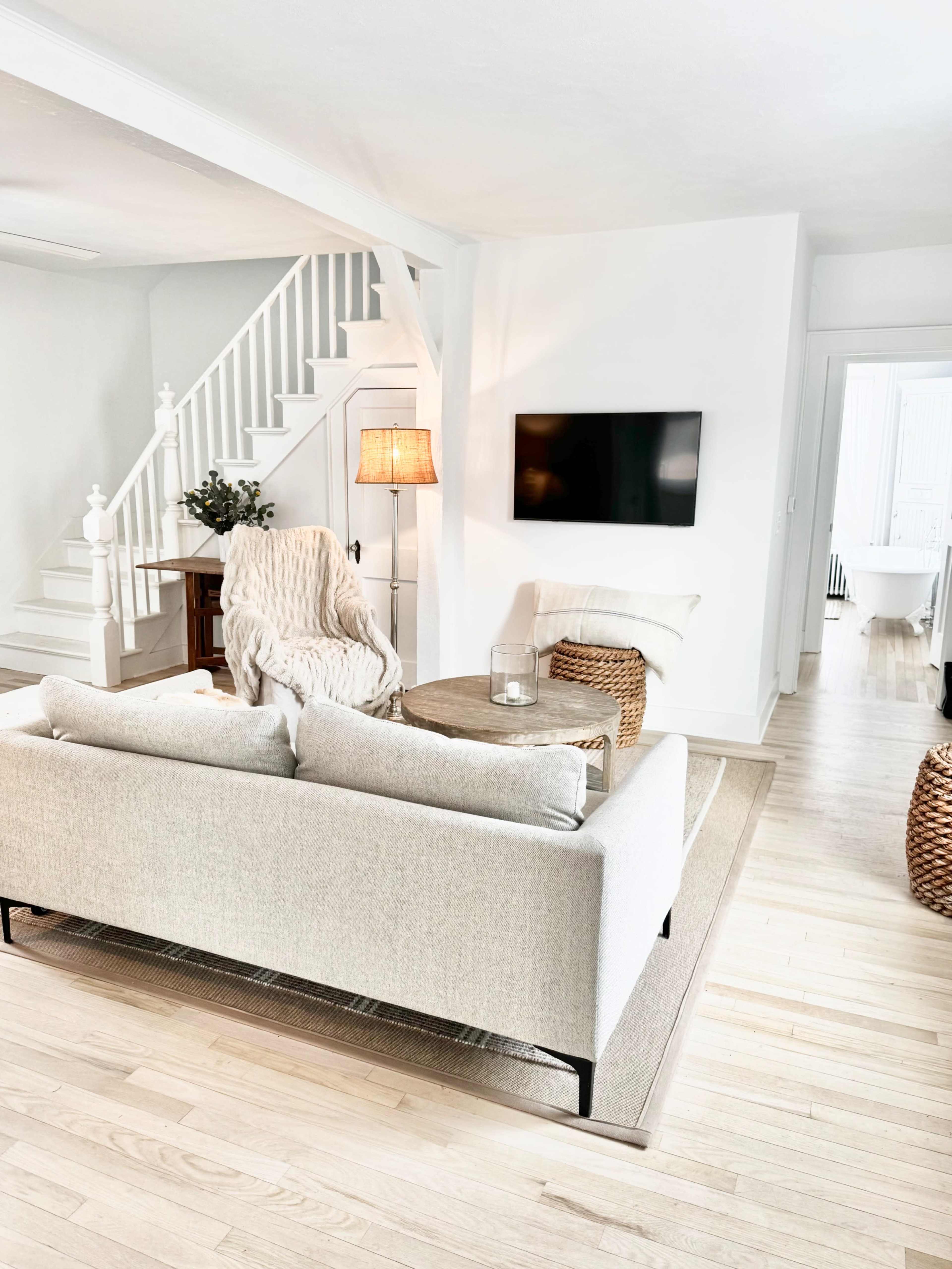 The image shows a bright, minimalistic living room featuring a light-colored sofa, a wooden coffee table, and a wall-mounted television, alongside a staircase and decorative plants.