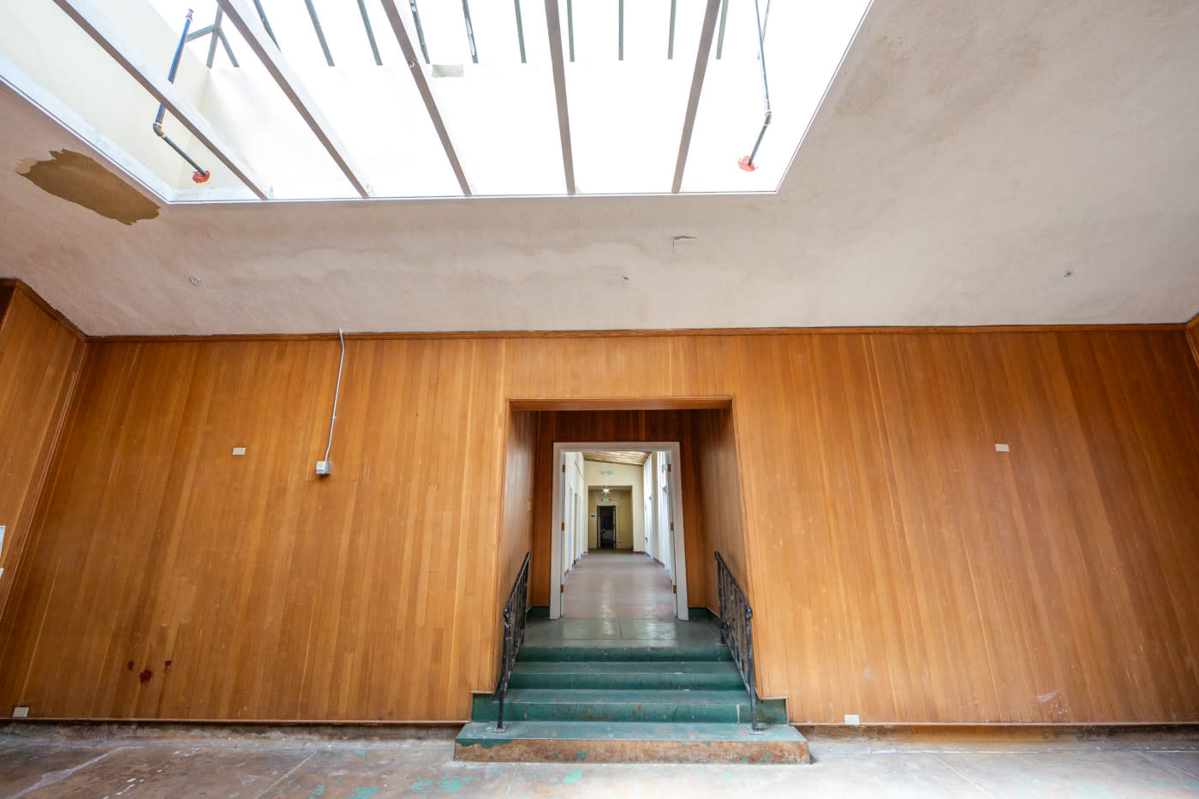 The image shows a large, empty hallway with wooden paneling and a set of stairs leading to another room, highlighted by natural light coming through a skylight.