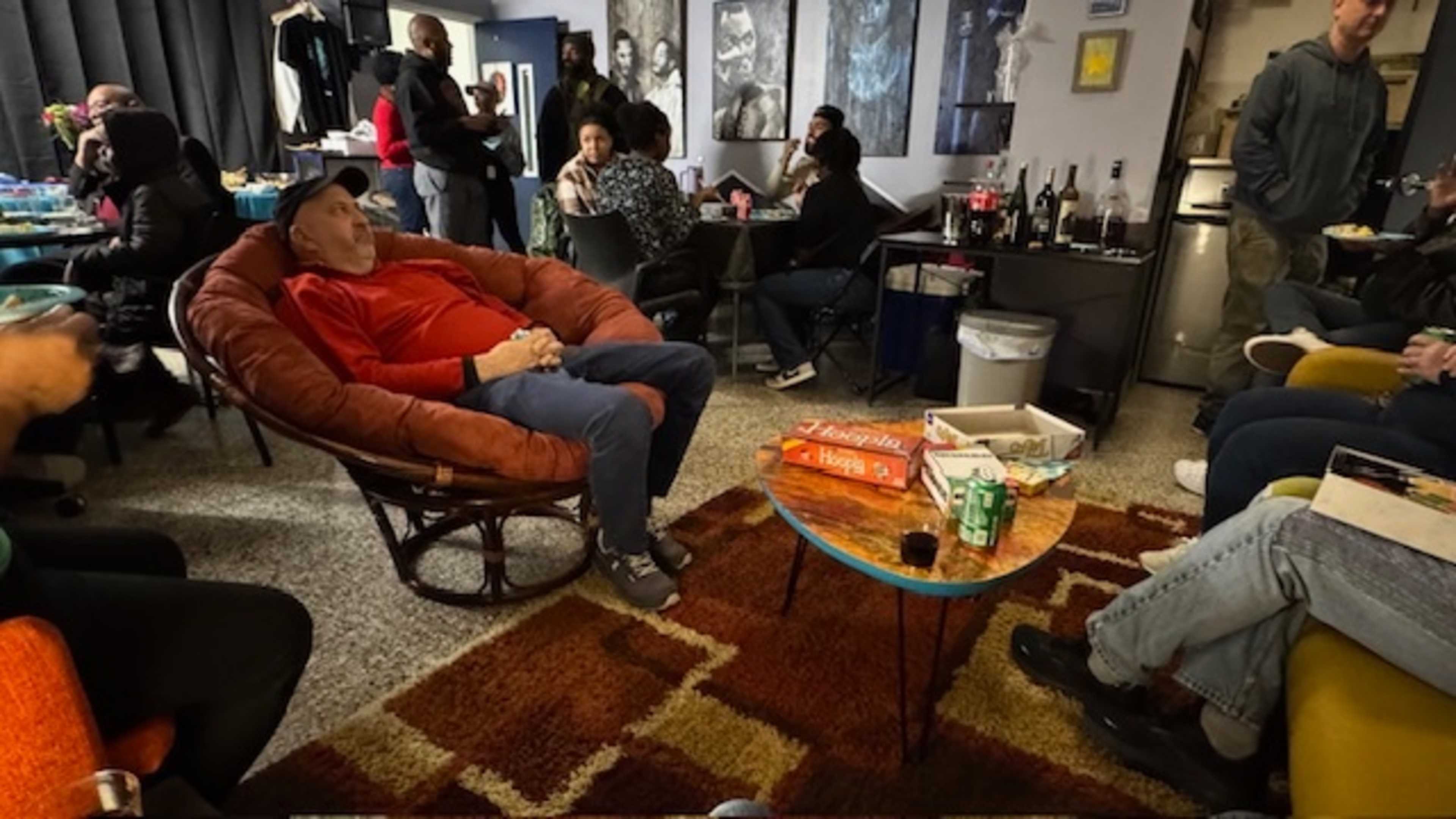 A group of people socializes in a cozy living room filled with chairs, a table with board games, and various bottles on a counter.