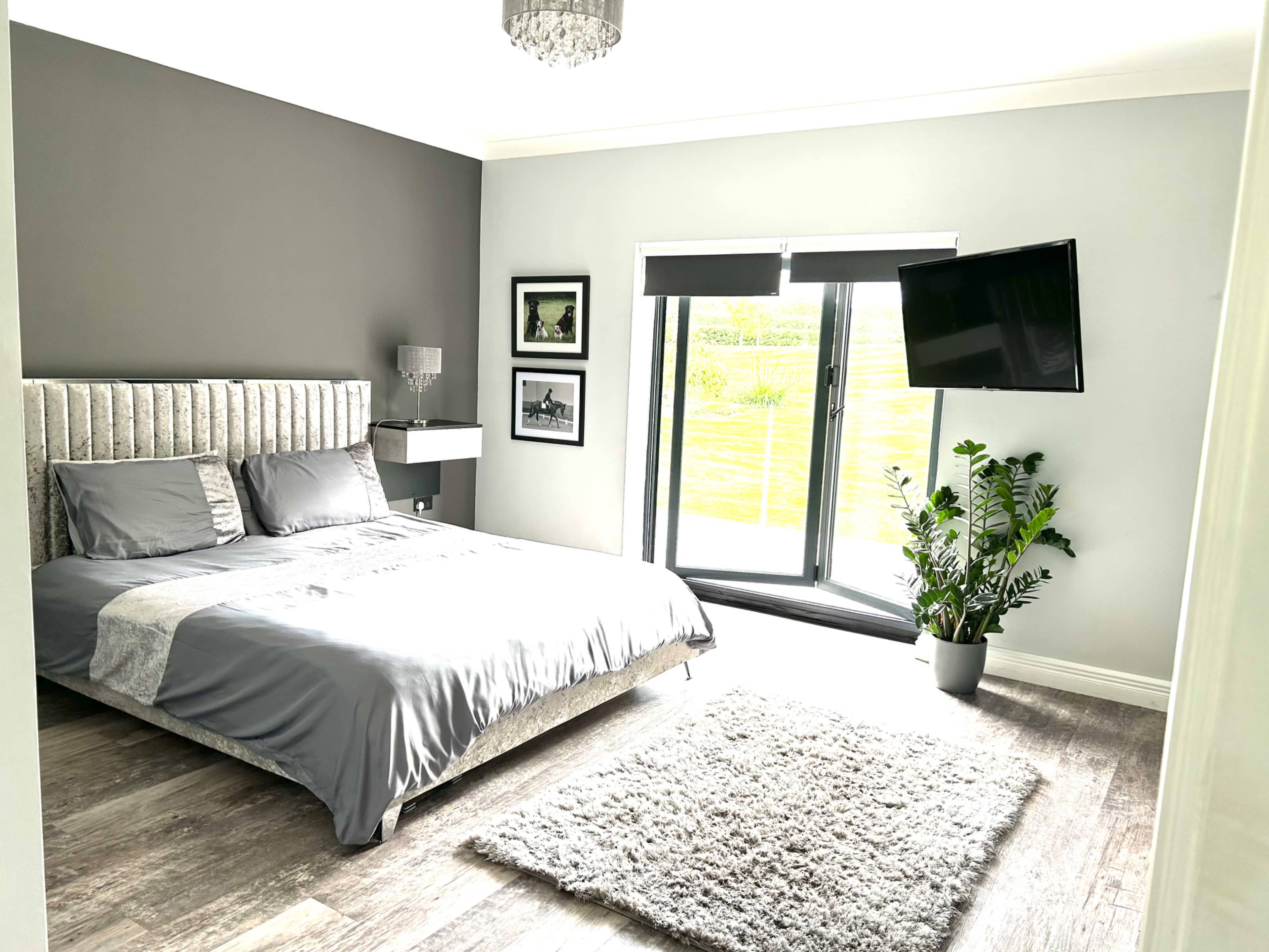 A modern bedroom features a bed with a gray headboard, a plush rug, a television mounted on the wall, and large sliding glass doors leading to an outdoor view.