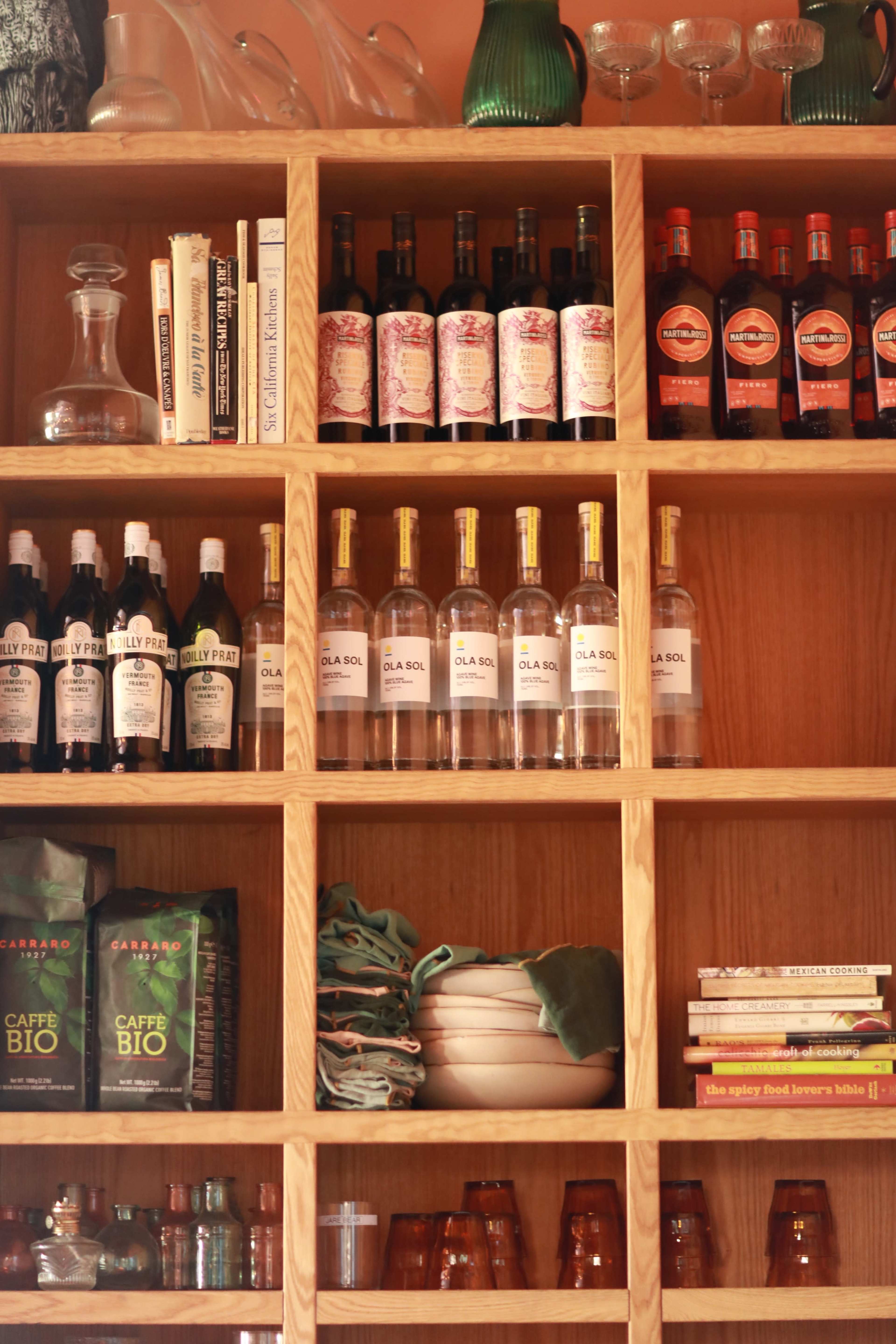 The image shows a wooden shelf filled with various bottles, books, and glassware organized neatly.