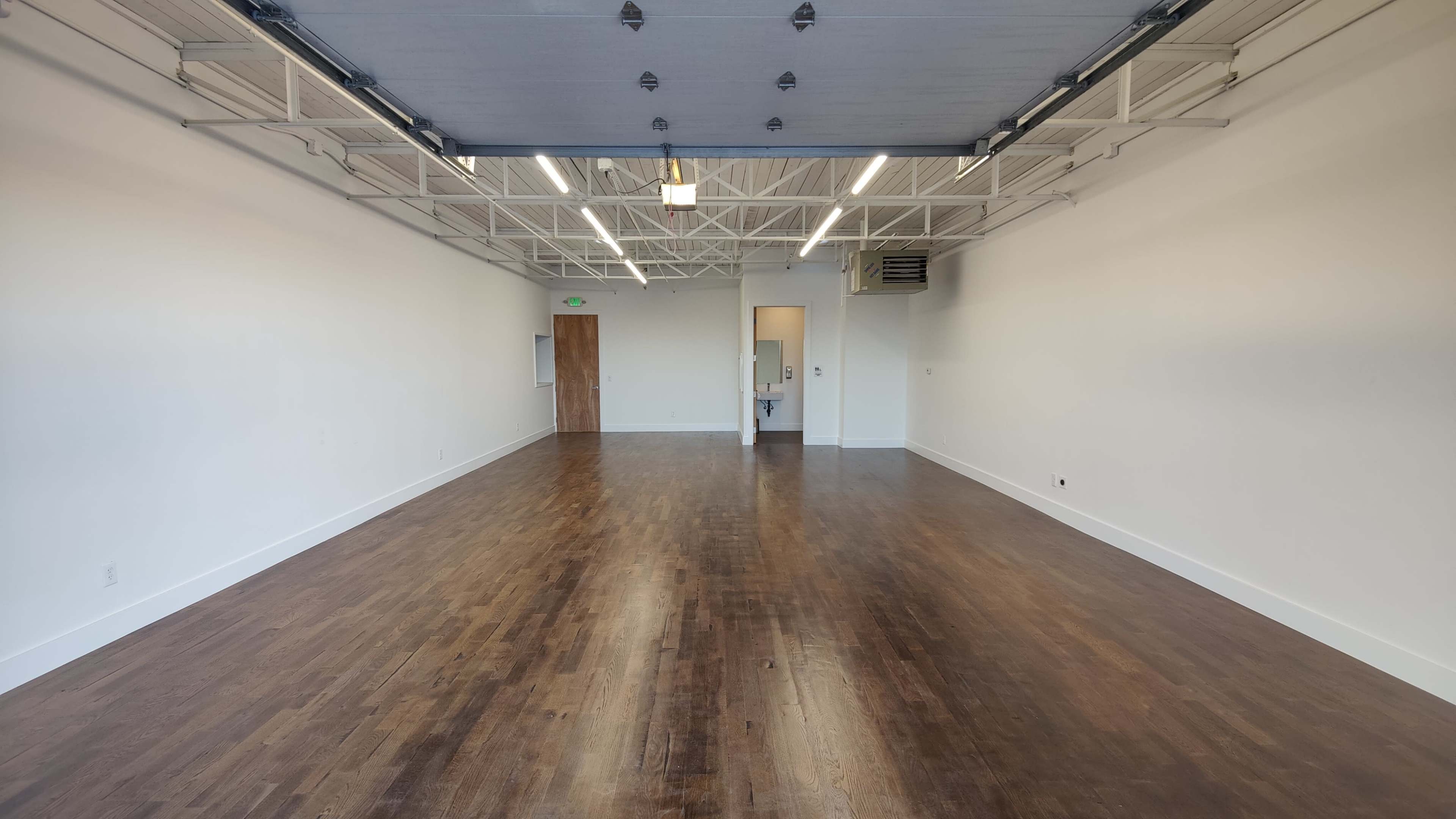 The image shows an empty room with wooden floors, white walls, and overhead lighting.