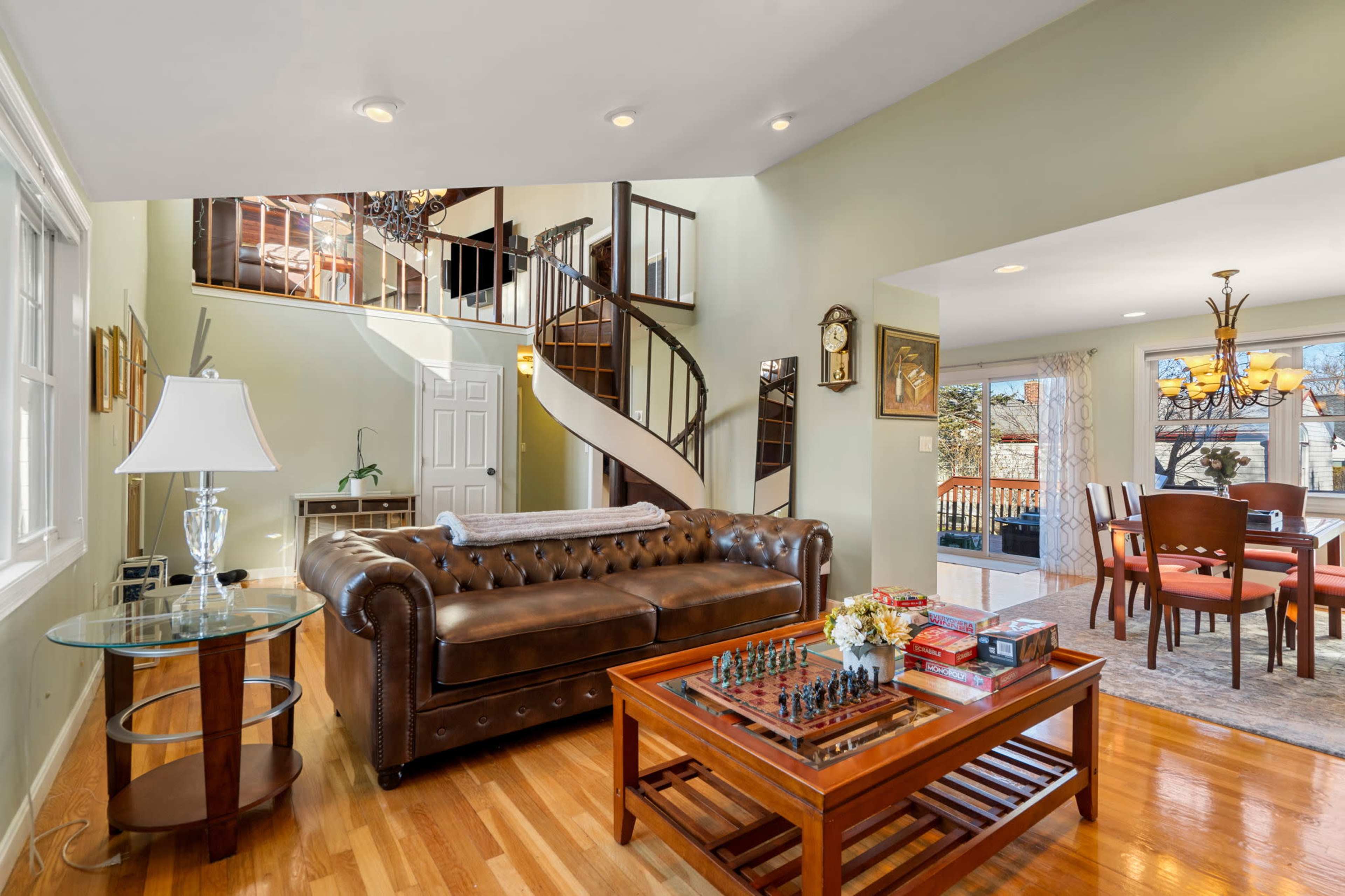 A living room with a leather chesterfield sofa, a wooden coffee table with a chess set on it, and a spiral staircase leading to the upper floor.