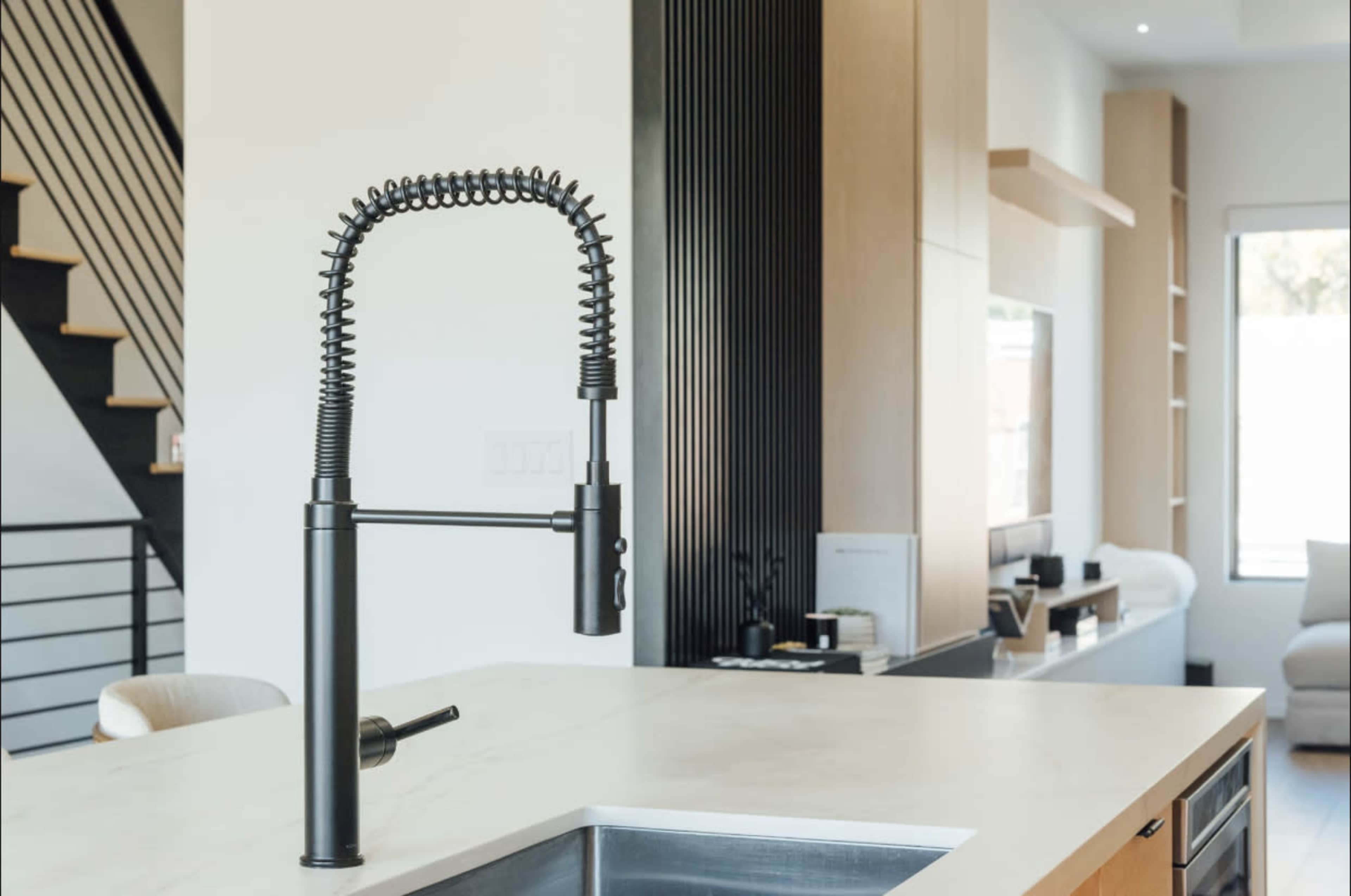 A modern kitchen features a black pull-down faucet on a marble countertop with a staircase and living area in the background.