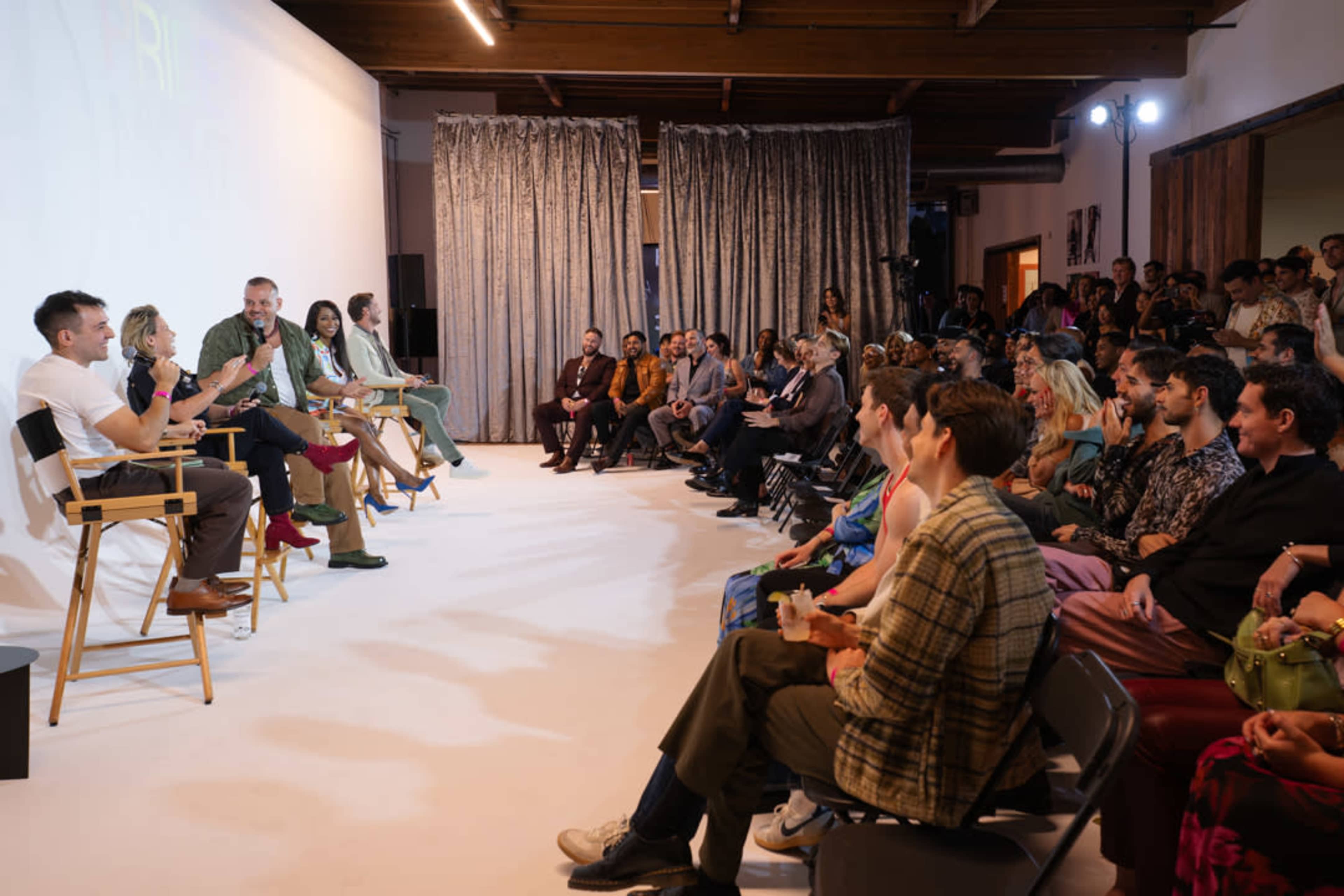 A panel discussion takes place on a stage in front of an audience seated in rows, with speakers on chairs engaging in conversation.
