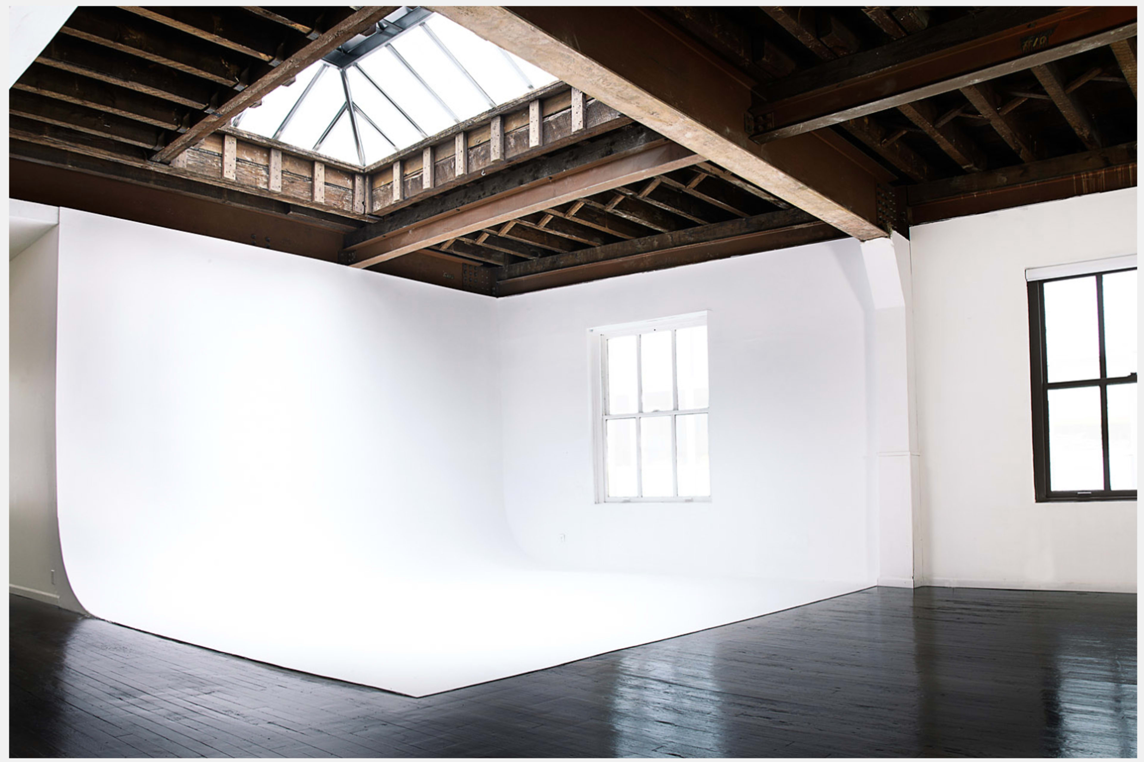 An empty photo studio with a seamless white backdrop, large windows, and exposed wooden beams.